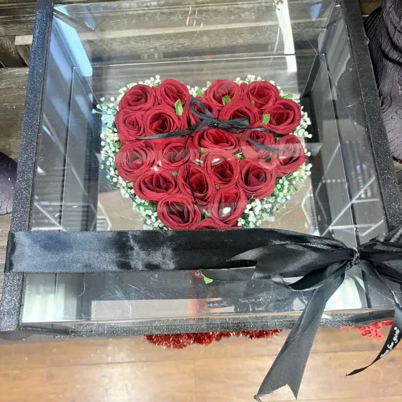 Heart Box With Roses - Tightly packed red roses formed into a perfect heart create an immediate romantic impression - ideal as a birthday gift for a partner, an anniversary bouquet, or a Valentine's Day surprise. The heart-shaped presentation reads as intentional and intimate, so the person you're thinking of will feel specially celebrated the moment the lid lifts.  The roses sit snugly inside a black gift box with a clear plastic lid, edged by a ring of delicate white filler blooms for crisp contrast against the deep crimson center. A black ribbon and bow finish the polished, formal look; touches of green foliage peek through to soften the arrangement and add natural texture. The compact, decorative format is designed for display on a bedside table or desk and arrives ready to present.