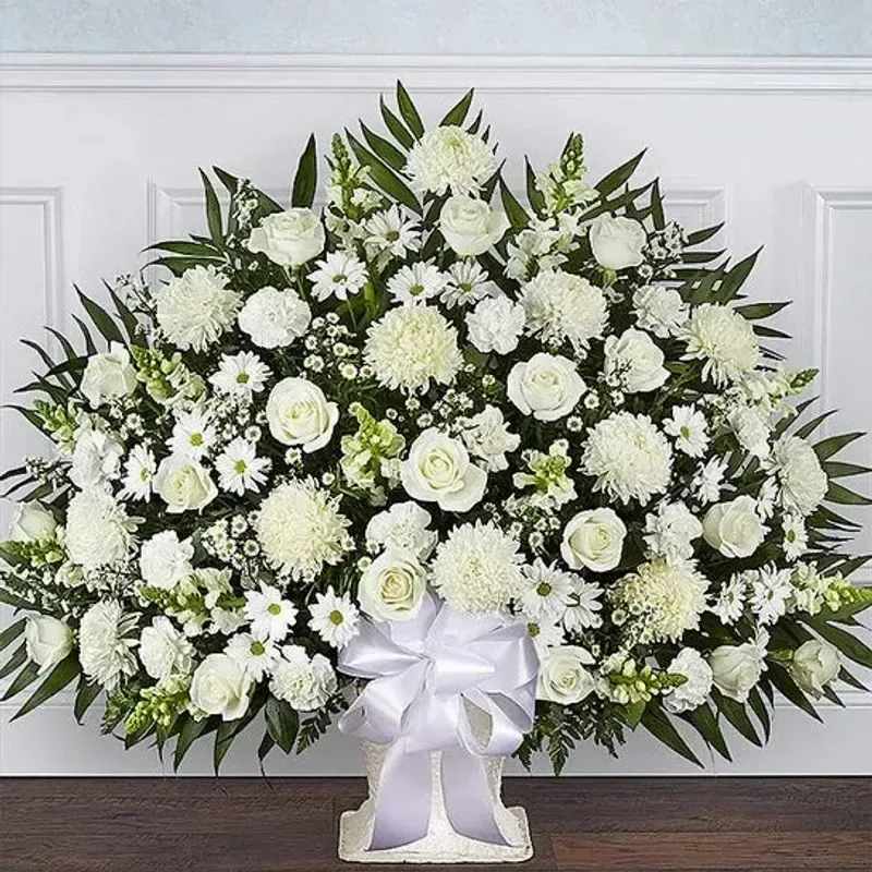 Purity In Love - A serene wall of white blooms-Purity In Love is a large standing spray arranged for sympathy and memorial services that will make the person you're honoring feel respectfully remembered. This funeral-style flower arrangement and sympathy tribute combines white roses, daisy-like blooms, chrysanthemum heads and snapdragons to create a calm, formal statement; the white-and-green palette reads as timeless and consoling for a graveside service or memorial ceremony.  The design is symmetrical and densely packed on a white floral foam base, framed by deep green foliage that heightens the crisp white flowers. A white ribbon bow at the base and a white standing easel complete the classic presentation; the overall scale is expansive to ensure visibility at viewings and services. Textures range from the soft rose petals to the rounded chrysanthemum centers and the upright snapdragon spikes, giving the piece visual depth while keeping the tone understated.  This arrangement is suited to express sympathy, comfort grieving family members, or mark a memorial milestone-it's the kind of piece that helps the recipient feel truly honored and seen. Care is simple: the foam base keeps stems hydrated for the duration of services; choose delivery to the funeral home or chapel for timely placement on arrival.