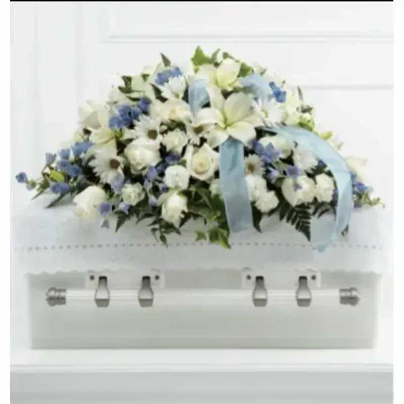 FTD® Tender Treasure Casket Spray™ - Soft white roses and pale blue delphinium create a calm, dignified statement for a sympathy or funeral casket spray that honors the life you're remembering. This large, broad arrangement reads as serene and traditional - a full, rounded profile laid across the top of a white casket, with clustered white blooms and small blue accents that convey quiet respect and gentle comfort to the person you're thinking of.  The design pairs roses, carnations, daisies and delphinium with touches of greenery for a cool, soft palette of white, blue and green. A light-blue ribbon is woven through the center and drapes over the flowers, adding a formal, memorial touch. Sized to cover the casket top, the low, wide shape keeps the focus on the flowers and the sentiment they carry, offering a tasteful, long-lasting tribute that will make family and friends feel comforted and honored.