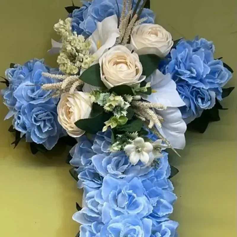 Memorial Wreath x15 - A devotional, cross-shaped sympathy wreath that offers a calm and reverent tribute for a funeral or memorial service. This memorial flower arrangement is composed of cool blue hydrangeas and ivory-white roses arranged densely to form a defined cross, creating an immediate visual of respect and remembrance for the person you're honoring.  The design pairs large blue hydrangea blooms with clusters of white roses and delicate white accent flowers, set against dark green foliage that outlines and strengthens the cross silhouette. The close-set flowers create a lush, textured surface with a tranquil blue, white, and green palette that reads formal and comforting from every angle.  Designed to convey solace and honor, this piece is suitable for church services, graveside committals, or as a home memorial. The arrangement is made to present well on an easel or casket and will maintain its composed appearance through typical service hours; please contact us for delivery and placement options to ensure it arrives where it will have the most meaning.