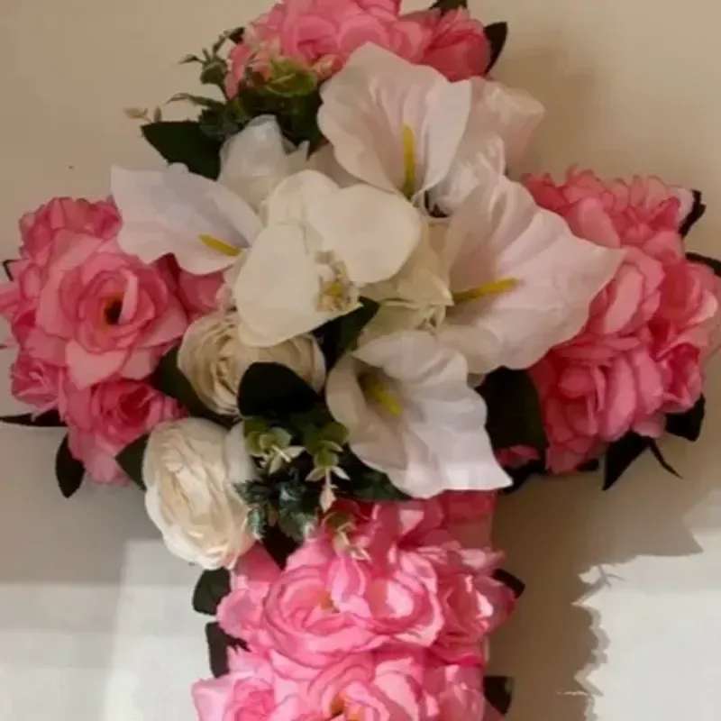 Memorial Wreath x13 - A formal cross-shaped sympathy wreath that offers a comforting, devotional presence for memorial and funeral services. The arrangement's bright pink roses frame the sides and lower section while large white calla lilies and white rose-like blooms fill the center, creating a calm, reverent focal point suitable for honoring a loved one.  Dense clusters of pink roses provide vibrant color and layered texture against the smooth, sculptural calla lilies and soft white blooms; touches of green foliage add fresh contrast and structure. The design is full, symmetrical, and intended to be displayed upright at a service or graveside to convey respect and remembrance.  Made to read clearly from a distance, this wreath is ideal for funerals, memorials, and religious observances where a dignified floral tribute is wanted. Please contact us for delivery details and placement guidance to ensure the piece arrives in perfect condition for the service.