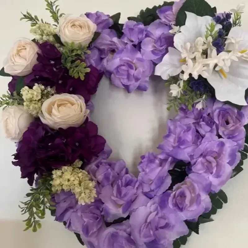 Memorial Wreaths X10 18" - A heartfelt heart-shaped wreath in soft lavender, deep plum and ivory tones designed for sympathy and funeral display - perfect as a memorial tribute that says "you are remembered" with quiet elegance. The romantic silhouette and gentle color palette make this arrangement especially fitting for honoring a loved one, a partner, or someone who cherished floral beauty.  The 18" wreath is densely clustered with purple blooms, cream and white roses, and small pale filler flowers for a plush, rounded look. Dark foliage frames the edges to define the heart shape and add contrast, while the mix of lavender, deep plum and pinkish highlights creates a layered, sentimental texture. The composition reads as both decorative and reverent, with the roses providing a focal point amid delicate accents.  Send this wreath to mark a funeral service, memorial table, or graveside tribute - the kind of piece that helps the recipient feel seen and supported. It arrives ready to display; for best longevity place out of direct sun and away from heat to preserve the flowers during services and viewings.