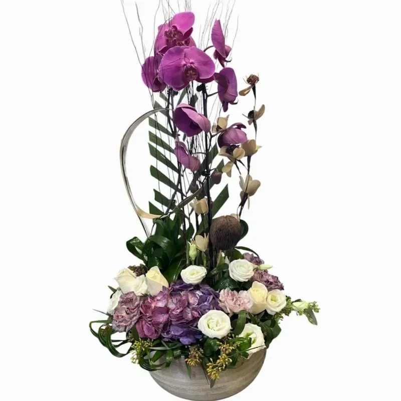 Purple craze - A tall cascade of purple Phalaenopsis orchids makes the first impression here - Purple Craze is a sculptural flower arrangement designed to delight for birthdays, anniversaries, thank-you gestures, or a chic housewarming gift. The vertical line of orchids draws the eye upward while a dense, rounded base of blooms grounds the design, so the person you're sending it to will feel both celebrated and thoughtfully seen.  At the base you'll find a plush cluster of white roses, lavender and pink hydrangea, and mixed greenery that softens the vivid orchids and adds texture. Decorative twigs and a curved metal ring arch above the composition for an artistic, contemporary silhouette, all arranged in a round ceramic vase (gray tone) with additional round container styling. The purple, white, pink, and green palette reads elegant and slightly dramatic without feeling overdone.  Perfect for anniversaries, Mother's Day, congratulations, or "just because" moments, this arrangement suits a refined home or office display and offers long visual impact thanks to the sturdy orchid stems and full hydrangea heads. Order with free delivery for a same-week surprise; to keep blooms fresh, place in cool indirect light and refresh vase water as needed.
