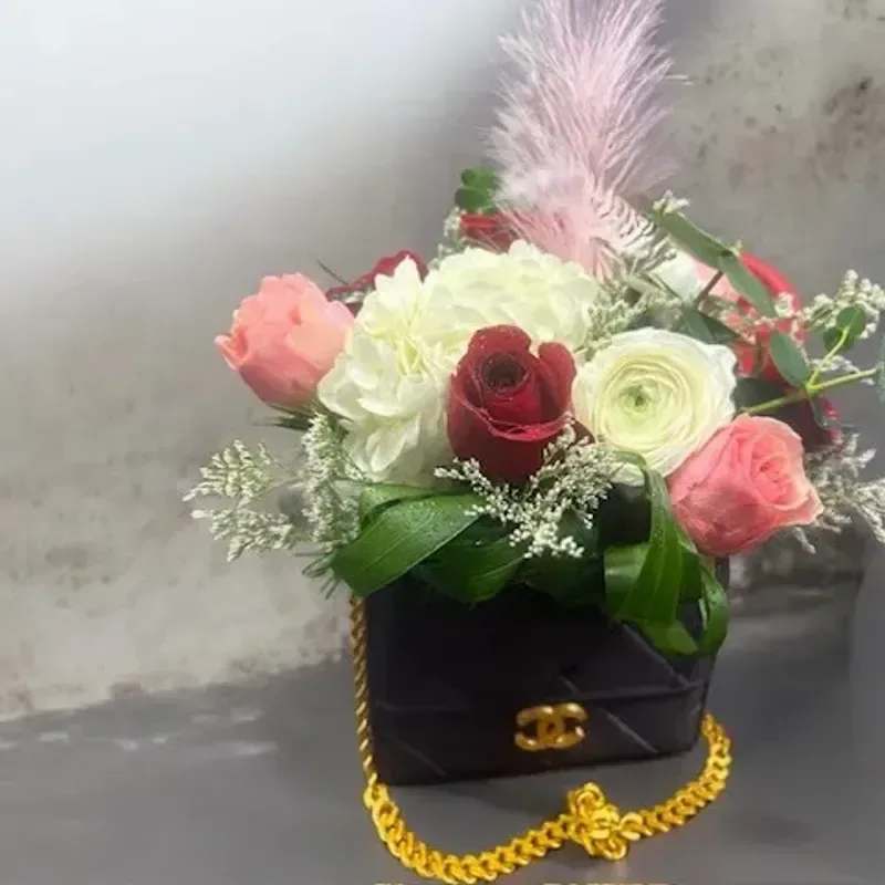 Luxury Inspired By Chanel - A compact black handbag-shaped box dressed with a gleaming gold chain and clasp holds a luxe mix of pink, white and deep-red blooms that reads like a fashion statement. This flower arrangement for gifting and special celebrations-think birthdays, anniversaries, or an elegant "thinking of you" surprise-makes the recipient feel effortlessly celebrated: the contrast of the black container against pastel and jewel tones creates an instant wow.  Inside the box are roses, hydrangea and ranunculus layered with airy white filler for texture and a pale pink feather accent for height and playful movement. The gold clasp and decorative feathers give a couture finishing touch, while the compact silhouette keeps it suitable for a bedside table or desk display. Ideal for someone who loves chic, fashion-inspired gifts, this arrangement arrives ready to display with no additional vase needed.