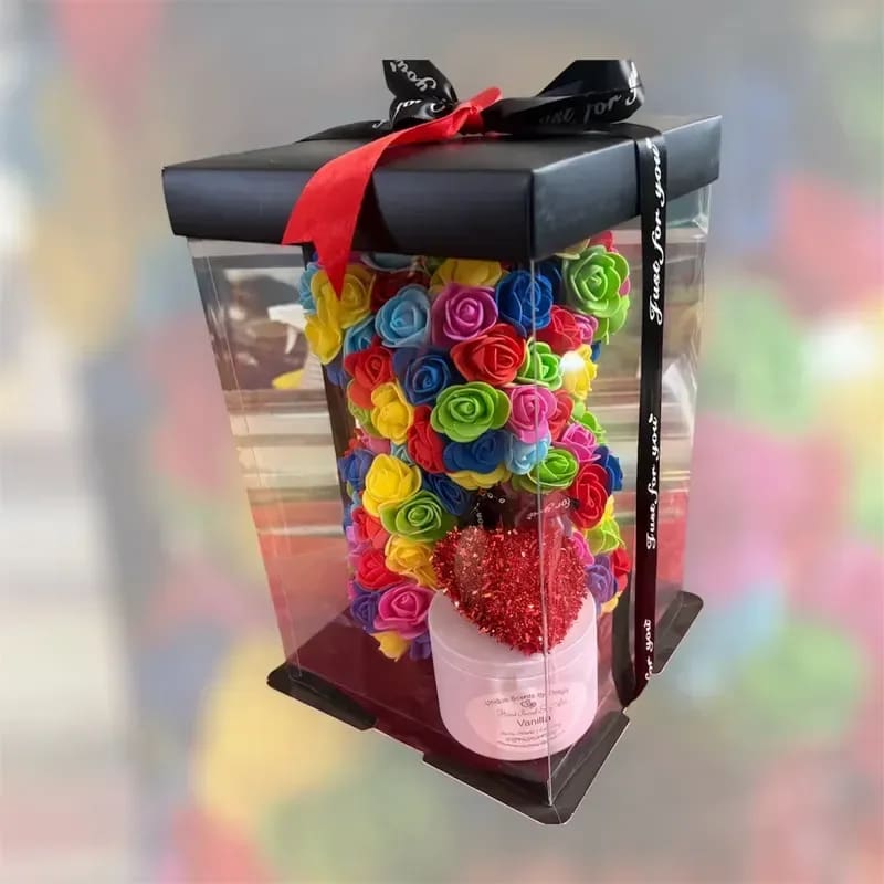 Multi Colour Rose Bear - A playful, candy-colored rose bear boxed for gifting-perfect as a birthday bouquet alternative or a just-because surprise. This flower arrangement features a dense cluster of small silk roses shaped into a charming bear form, with mixed colors including bright red, pink and playful blue that read cheerful and romantic. The boxed presentation makes it an ideal love-and-romance gift or a fun pick-me-up for someone who loves bold, joyful color.  The bear sits inside a clear rectangular plastic gift box with a contrasting black lid; a red ribbon crosses the top and a glittery red heart decoration at the front adds an extra touch of whimsy. A secondary black ribbon and a pink container inside complete the layered look, giving the arrangement polish and ready-to-display appeal. Because the blooms are high-quality silk, the recipient gets a long-lasting keepsake that looks fresh without watering or special care-simply unwrap and place on a shelf or bedside table to brighten their day.