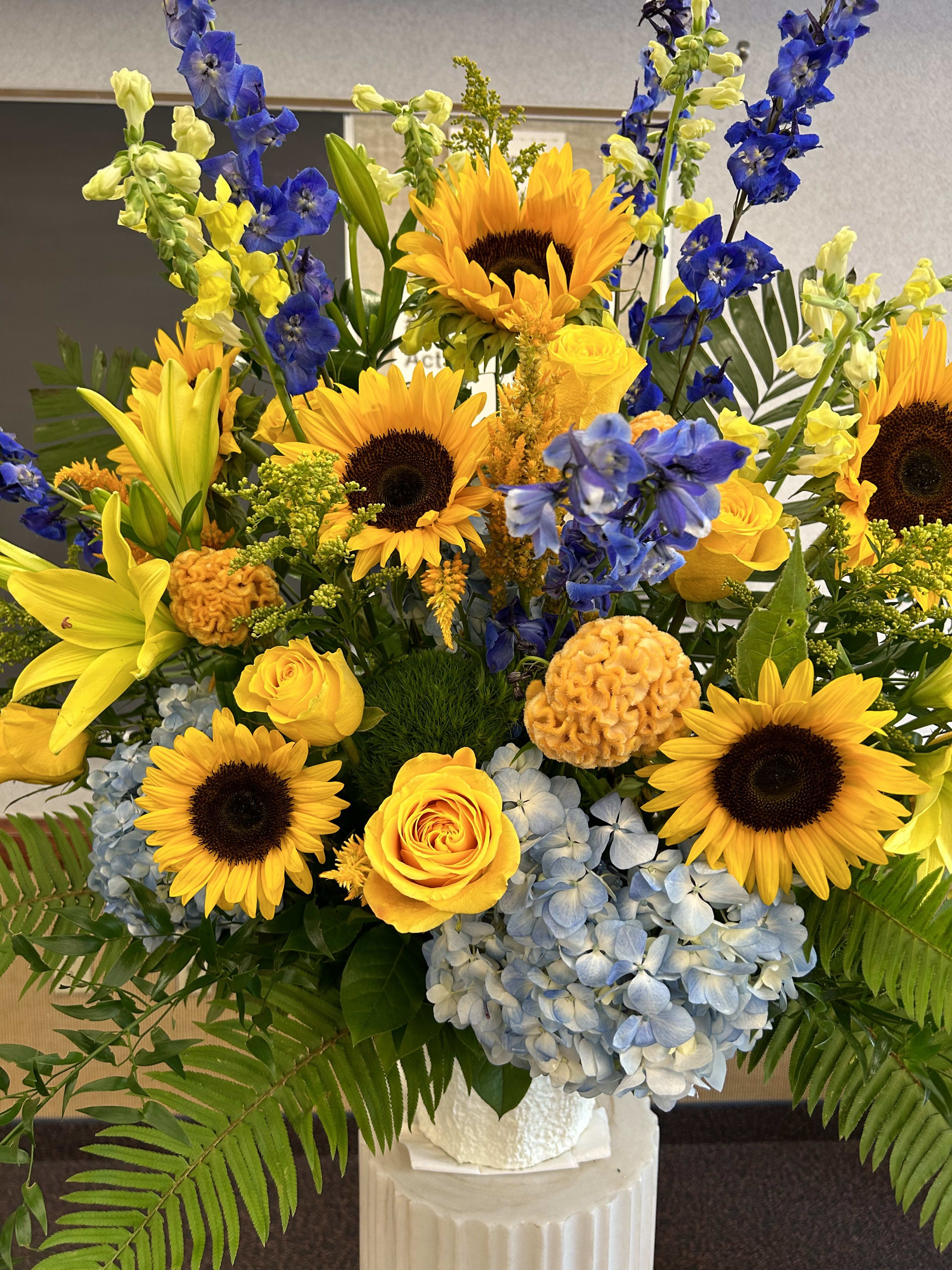 Bright bouquet of sunflowers, yellow roses, blue hydrangeas, and lilies in a white vase