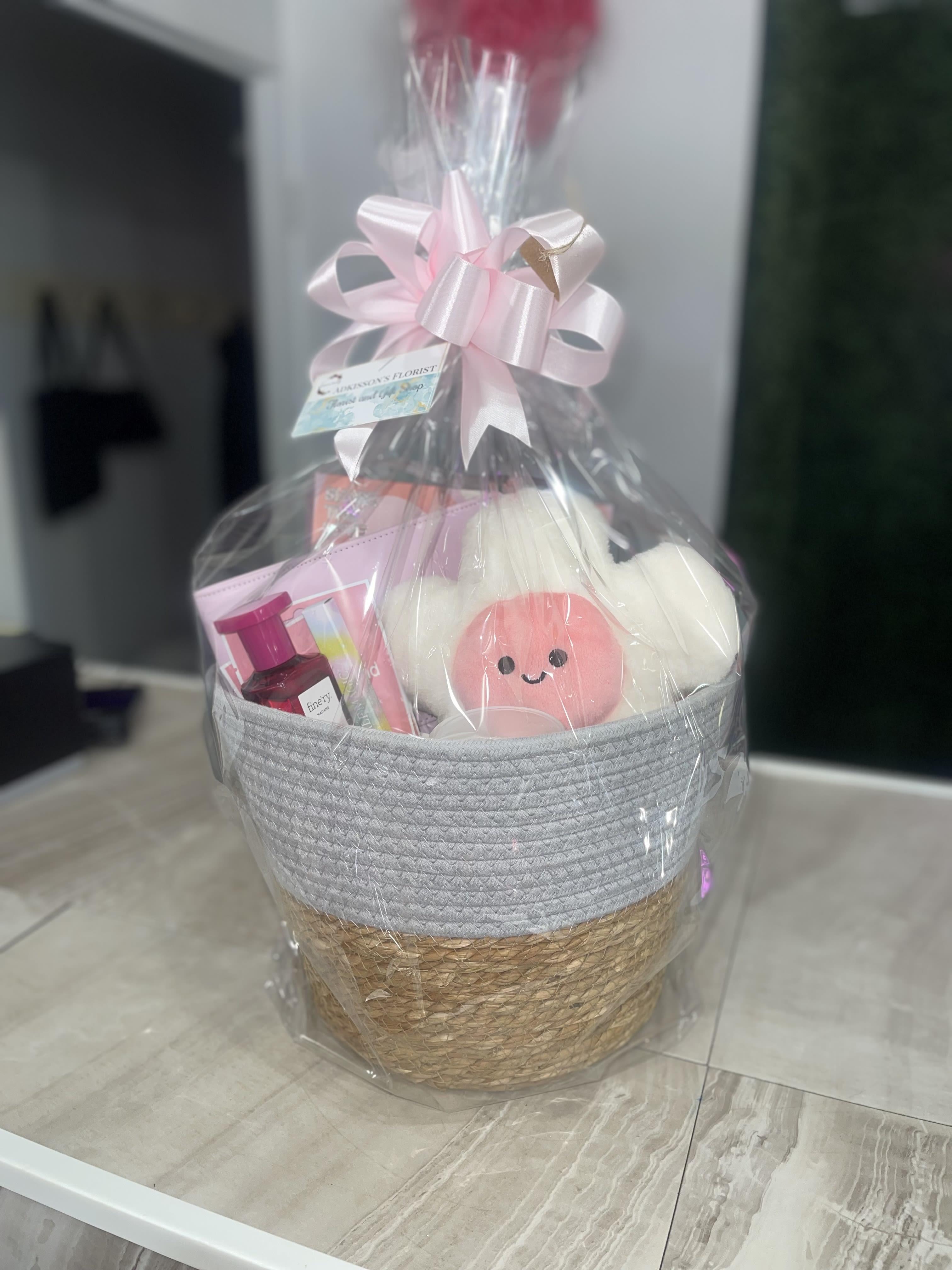 Gifts For Her - Make this gift basket a heartfelt pick for birthdays, thank-you gifts, or a get-well surprise. The presentation reads like a thoughtful "gifts for her" set-wrapped in clear cellophane and finished with a large pale pink ribbon bow-so the person you're thinking of will feel cherished the moment it arrives.  Inside a woven basket a smiling plush that sits among pink-toned gift items and a pink bottle, all arranged closely to create a cheerful, feminine palette. The package includes a product tag and is tied with pink satin ribbon, giving a handmade, boutique feel that's easy to display right out of the wrap.  Perfect for someone who appreciates cute keepsakes and gentle colorways, this basket sends a message of warmth and care without being formal. The clear wrapping and full bow keep contents secure for delivery; no special care is needed-simply present as-is for an instantly joyful gift.