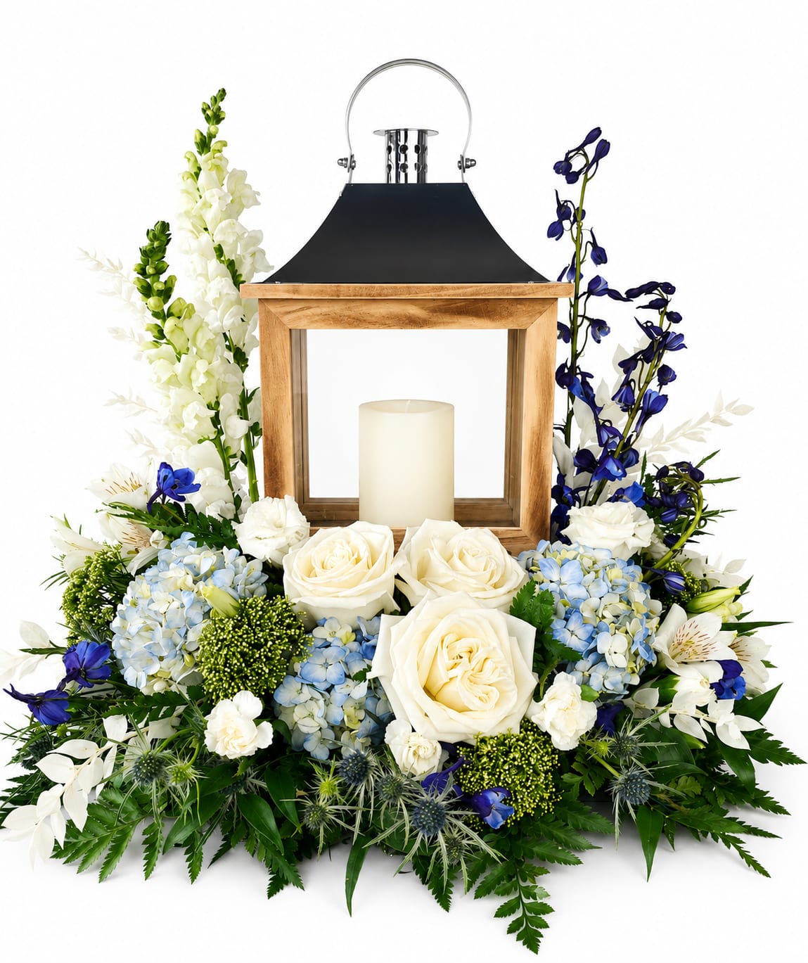 Lantern Floral - A cool, serene centerpiece built around a rustic wooden lantern creates a quiet, elegant statement-ideal as a sympathy arrangement or for a memorial service. The lantern floral arrangement combines white roses and creamy hydrangeas with pale blue and deeper blue blooms plus tall white snapdragon spikes to produce a layered, formal look that reads both polished and peaceful. The included pillar candle in a glass holder softens the scene, giving the person you're honoring a gentle, steady glow.  This low, wide composition sits balanced and symmetrical, with taller floral accents on each side to create ceremonial presence without towering over a table. Textures range from the lush roundness of roses and hydrangeas to the vertical snapdragons and delicate alstroemeria, all in a cool palette of white, blue, &amp; green. ( Lanterns Styles and colors may vary on availability)  Choose this piece to offer comfort and convey respect-the kind of arrangement that helps someone feel seen and remembered during a difficult time. Arrives ready to display on a table or altar; keep blooms away from direct heat and replace the candle if you plan extended use.