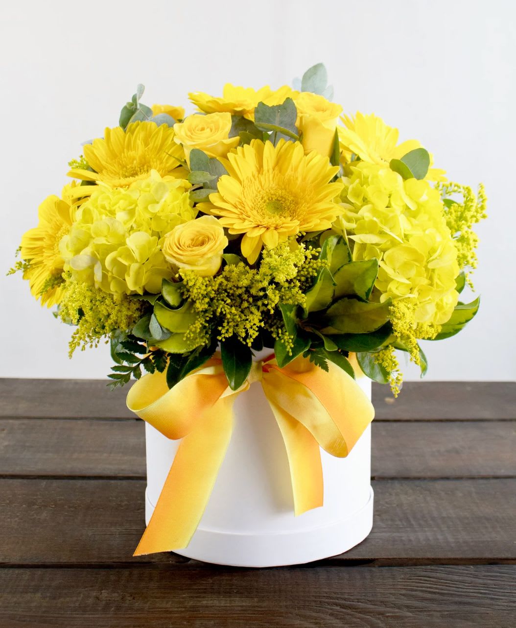 Sunny Delight Floral Hatbox - Brighten any space with the radiant charm of our Sunny Delight Floral Hatbox. This stunning arrangement bursts with a joyful medley of fresh yellow blooms, including vibrant gerbera daisies, classic roses, hydrangeas, and delicate sprigs of filler flowers, beautifully complemented by lush greenery and silver dollar eucalyptus leaves. Presented in a sleek white hatbox and tied with a cheerful ombré yellow ribbon, this bouquet is perfect for celebrating special occasions or simply spreading sunshine.  Perfect for: Birthdays, congratulations, get well wishes, housewarmings, or to bring a smile to someone’s day.  Handcrafted with care and available for same-day delivery.