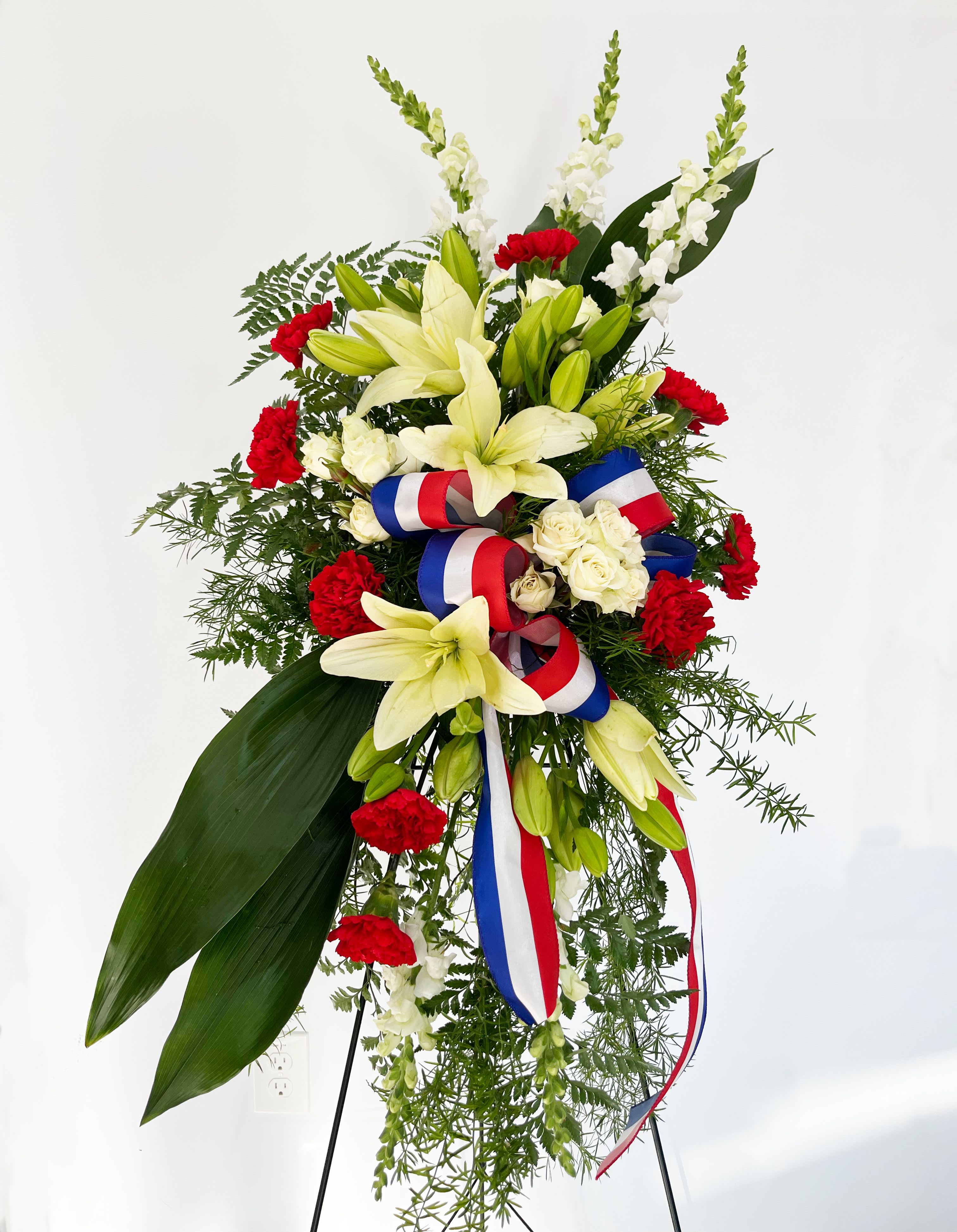 Stars of Life Standing Spray - STARS OF LIFE STANDING SPRAY  Share your respects with flowers that evoke the true spirit of a hero. Our Stars of Life spray is accented with a patriotic ribbon and features a gorgeous yet timeless collection of carnations, snapdragons, lilies and roses.  Please Note: The bouquet pictured reflects our original design for this product. While we always try to follow the color palette, we may replace stems to deliver the freshest bouquet possible.
