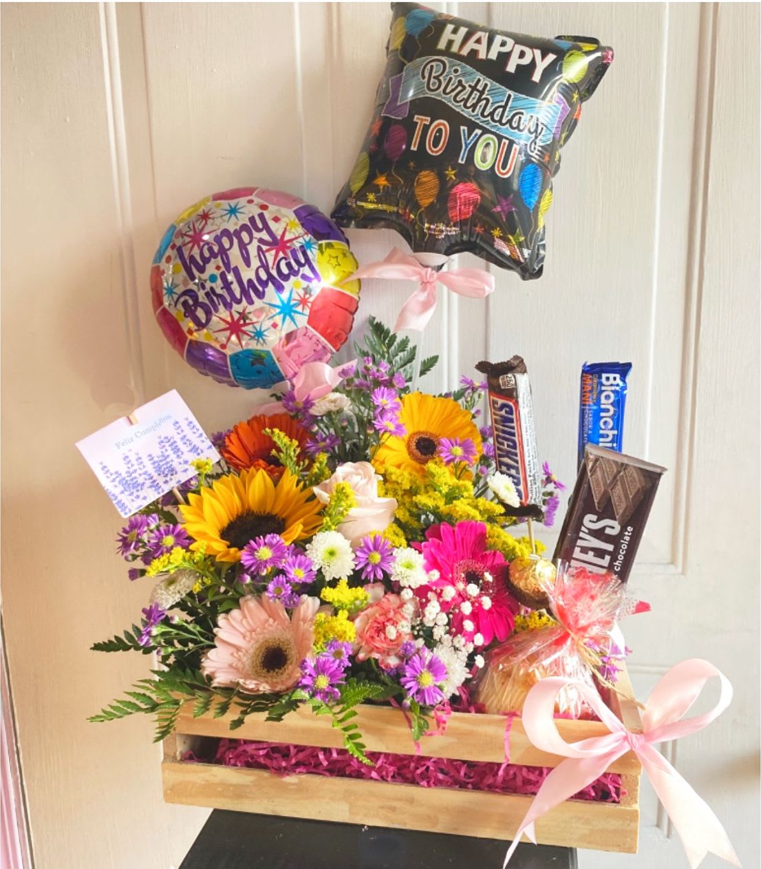 Birthday Celebration Box - Birthday Celebration Box with Sunflowers, Gerbera Daisies, Spray, Mini Carnations and Cushion Pom with accents of Purple Montecasino and Solidago. Includes 3 chocolate bars, A Vanilla Cupcake From ENTICING ICINGZ CAKE SHOP, 2 Small Happy Birthday Balloons. **BOX DESIGN WILL VARY *** Depending of the availability of flowers and material, substitutions are necessaries. 