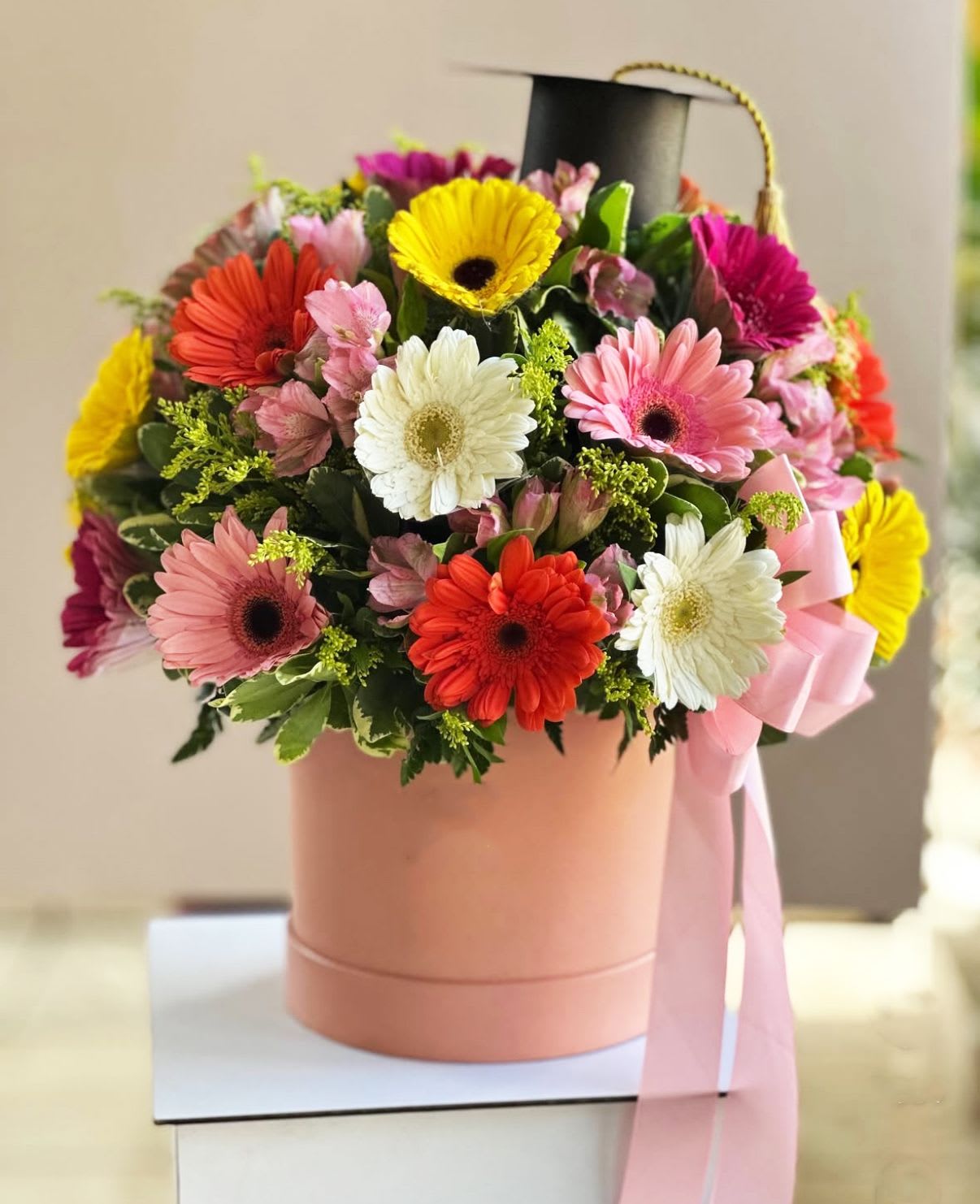 Graduation Gerbera Box - Celebrate life’s bright milestones with our Graduation Gerbera Box! This joyful arrangement features a vibrant mix of Gerbera Daisies and Alstroemeria, carefully hand-arranged in a soft blush hatbox and topped with a mini graduation cap — perfect for commemorating a proud achievement.