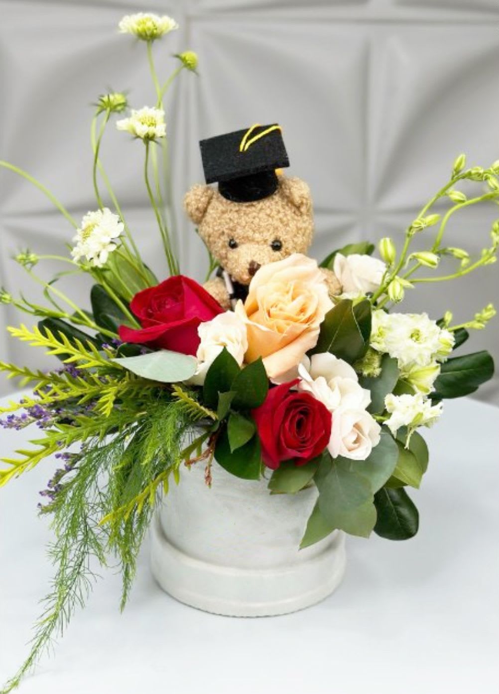 Smart Bear Blooms - Celebrate a special graduation moment with Smart Bear Blooms — a charming arrangement bursting with personality and elegance. Nestled in a sleek box, this design features a plush teddy bear proudly donning a graduation cap, surrounded by a lush mix of classic blooms and fresh greenery.