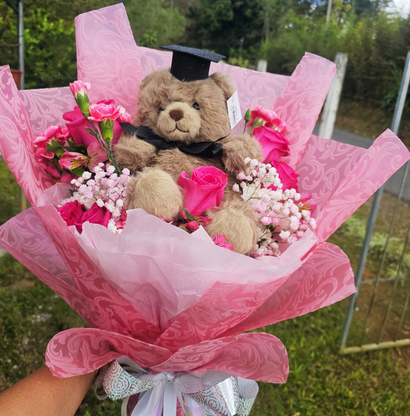 Pink Pride Graduation Bouquet - Celebrate success in style with the Pink Pride Graduation Bouquet — a charming mix of vibrant pink blooms, delicate baby's breath, and an adorable teddy bear in a graduation cap. The perfect gift to honor achievement with a hug and a smile.