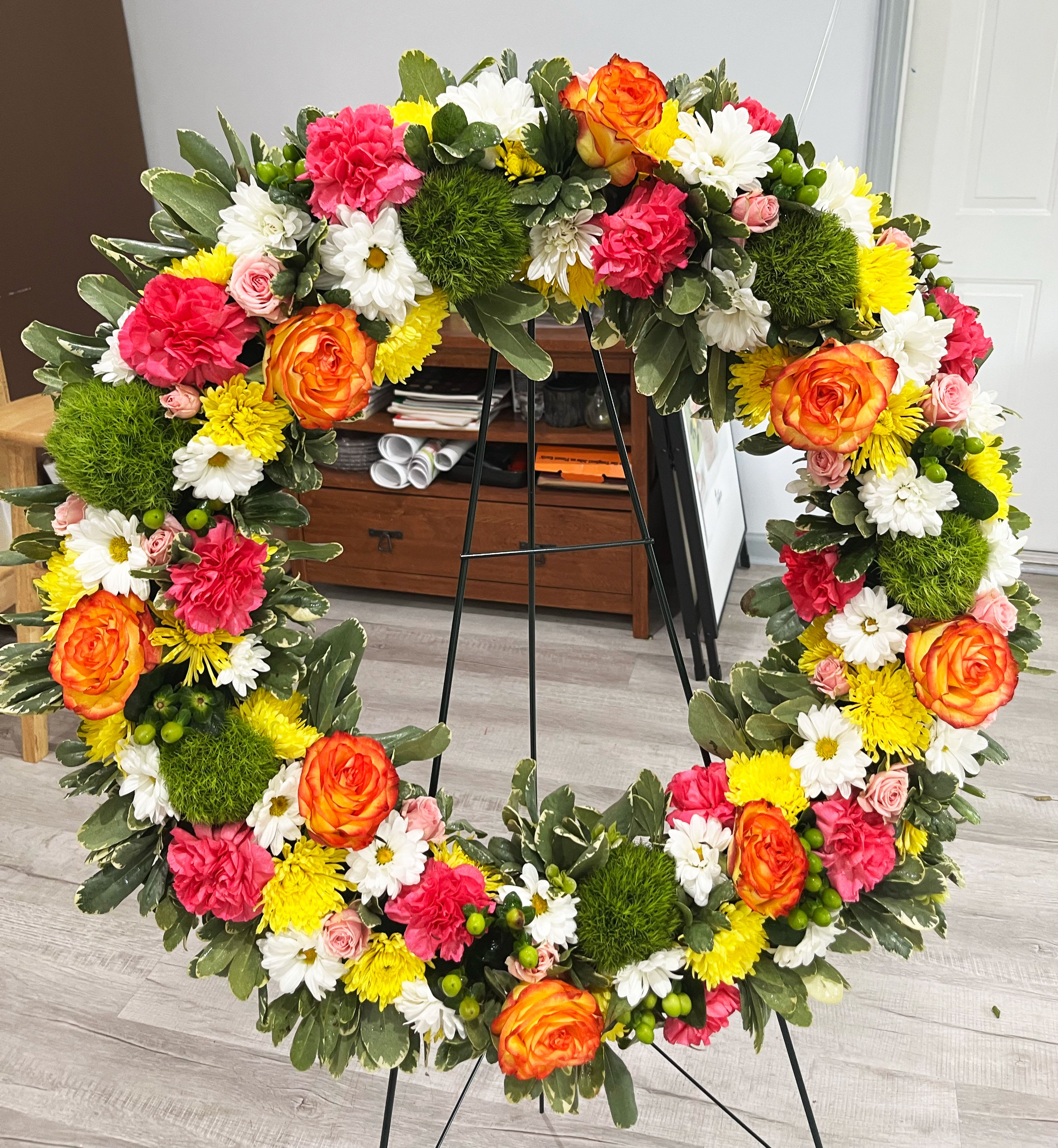 18" Radiant Tribute Wreath - Celebrate a life lived with joy and color. The Radiant Tribute Wreath is a vivid and uplifting sympathy arrangement featuring vibrant orange roses, hot pink carnations, sunny yellow chrysanthemums, white daisies, green button poms, and soft pink spray roses, all set against lush foliage. This standing wreath is a beautiful way to express love and remembrance with warmth and hope.