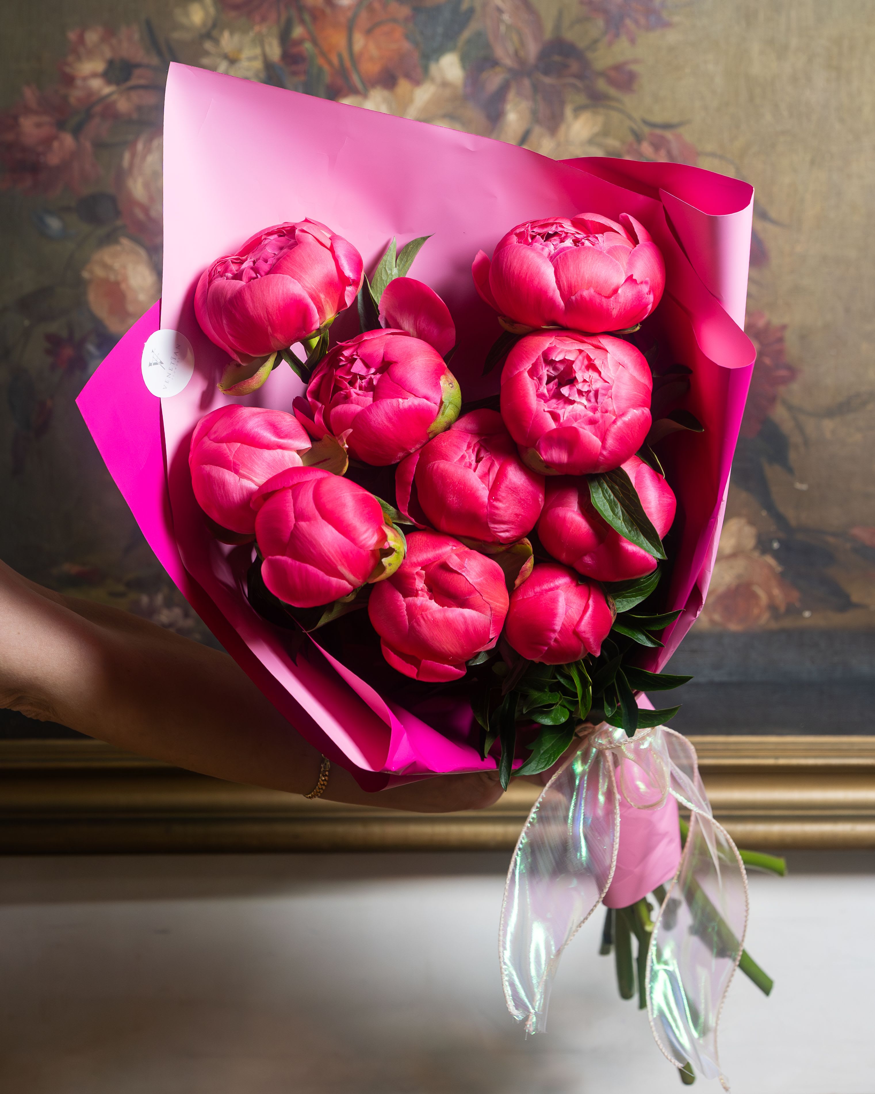 Coral Sunset Peonies Wrapped Bouquet - A lush cluster of vivid pink peonies makes a joyful, romantic statement-perfect as a birthday bouquet, anniversary gift, or Mother's Day surprise. This peonies wrapped bouquet puts the flowers and the recipient front and center: the layered, rounded blooms read as generous and celebratory, the saturated pinks feel distinctly feminine and cheerful, and the few dark green leaves that peek through add just enough contrast to keep the arrangement fresh and textured.  Each bouquet features tightly clustered peony blooms in various stages-from tight buds to fully opened flowers-wrapped in bright pink paper and secured with a sheer translucent ribbon and a round sticker label for a polished, gift-ready look. The wrapped presentation (pink floral wrap and additional pink paper) makes it easy to carry and give immediately; size feels lush and abundant without needing a vase, though the stems are vase-ready if you want to display them at home.  Designed to make the person you're thinking of feel truly seen and celebrated, this arrangement suits everyday gifting moments as well as milestone occasions. To keep peonies looking their best, place in fresh water upon arrival and trim stems slightly; ordering options and delivery timing will help ensure blooms arrive at their peak.