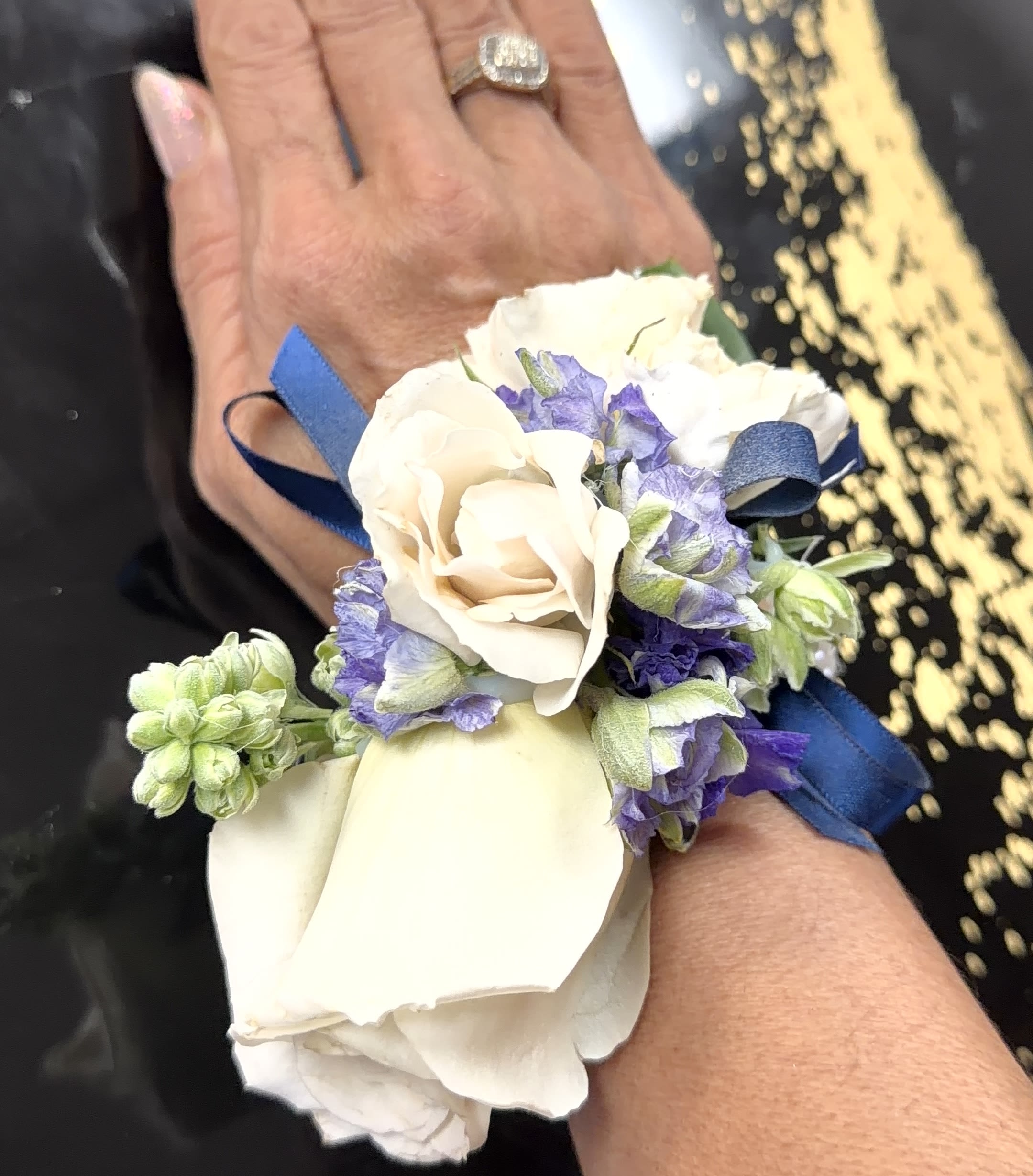 Blue. and white Corsage and Botonier - A cool-toned wrist corsage and matching boutonniere set designed for prom, wedding, or any special-occasion accessory that calls for refined elegance. The corsage centers on a layered white rose surrounded by white alstroemeria and delicate purple accents, so the person wearing it will look polished and thoughtfully dressed for the ceremony or reception. The boutonniere mirrors that same crisp palette, making this pair a coordinated choice for couples, attendants, or a host and their guest.  This compact arrangement combines white roses, alstroemeria, small green buds and leaves, and subtle purple florals to create a rounded, feminine silhouette. Hard goods include a blue ribbon tied around the corsage's wrist band, a white satin ribbon, and a strand of pearl beads for a polished finish; the boutonniere is sized to sit neatly on a lapel. The cool blues, whites, and soft purples give a fresh, formal look without overwhelming the wearer's outfit.  Perfect for bridesmaids, prom dates, mothers of the couple, or anyone who should feel special and coordinated-this set signals care and attention to detail. Order with your event in mind; the compact wristband and boutonniere are ready to wear and best kept cool and dry until the moment they're pinned or tied on for lasting freshness.
