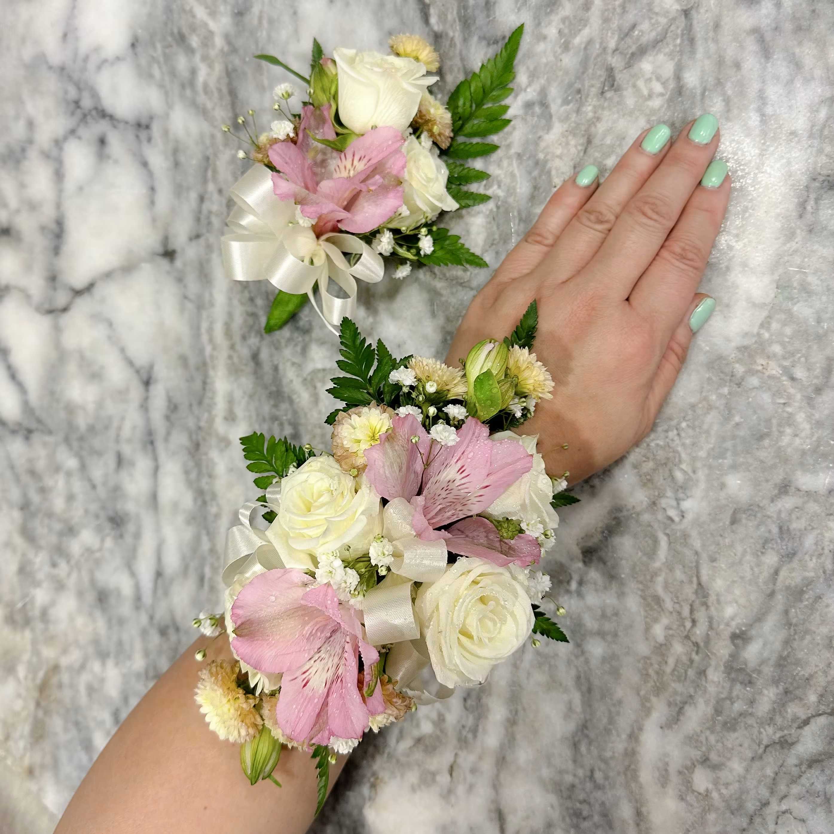 Corsage and Botonier pink - A rounded bouquet of soft pink and white roses framed with pale purple carnations and white alstroemeria creates a gently romantic centerpiece perfect for a wedding bouquet or a sentimental anniversary gift. This mixed-color floral arrangement and its matching smaller piece give the impression of a coordinated set-ideal when you want the person you're thinking of to feel celebrated and seen-with airy baby's breath and broad eucalyptus-like foliage lending a fresh, garden-style softness.  The arrangement is presented in clear glass vases (one larger, one small square) tied with satin ribbon for an elegant finish. Alongside the bouquets, two matching wrist corsages feature white roses, pink alstroemeria, petite daisy-like filler flowers, and bright green accent buds, each secured with ivory ribbon bows to sit comfortably on the wrist. The palette of pink, white, green and touches of purple reads delicate and feminine, making this set a graceful choice for weddings, prom, or any special occasion that calls for understated romance.  Give this coordinated set when you want her to feel truly noticed-whether walking down the aisle, stepping into prom night, or marking an intimate anniversary. Vases are simple clear glass for easy placement on a table; corsages are scaled for wear and arrive ribbon-secured for easy fastening. For best longevity, keep flowers in cool, draft-free conditions and refresh the water daily.