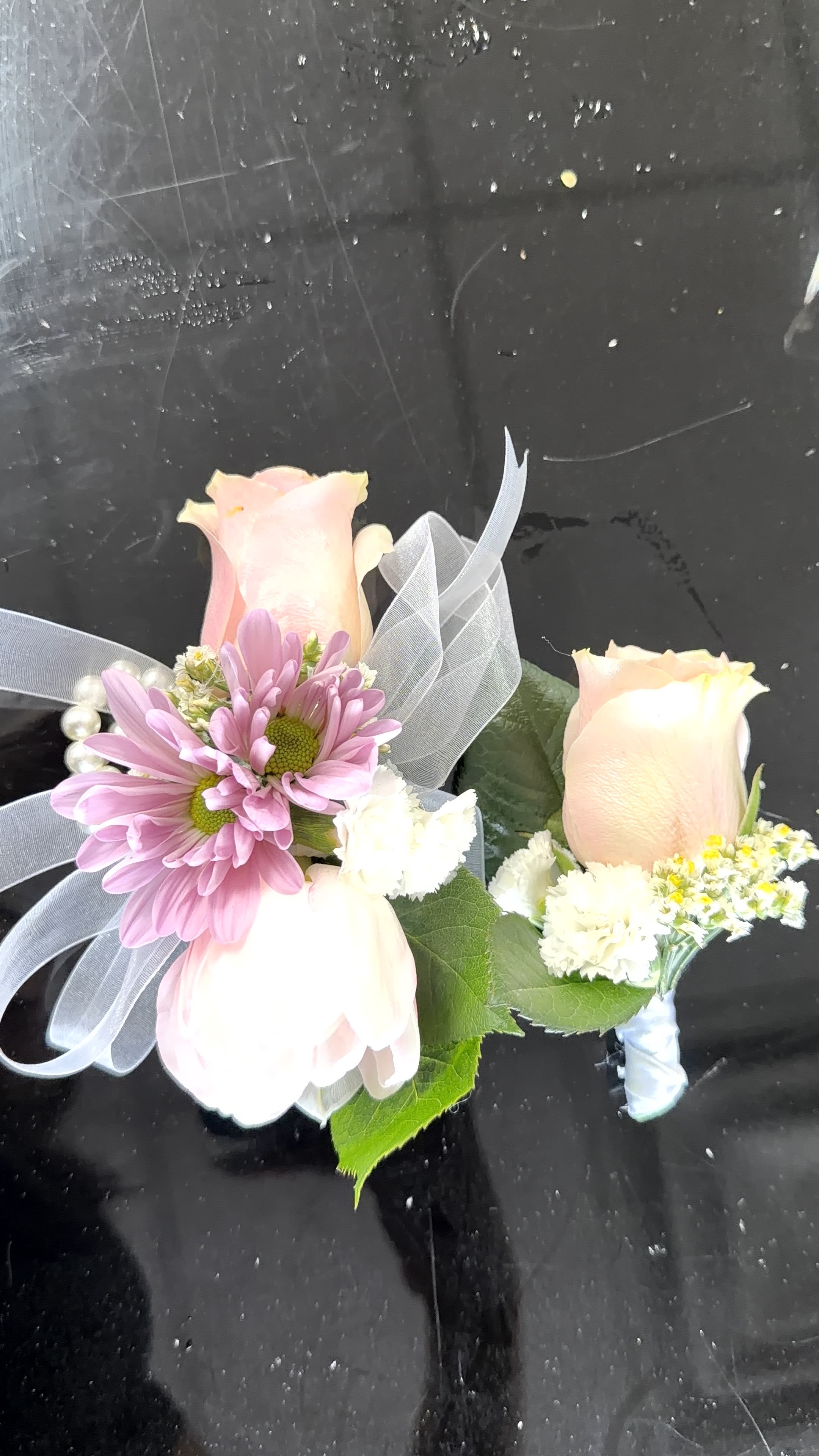 Pink Corsage and Botonier - Pale pink and peach roses paired with lavender-pink daisies make these wearable floral accents feel quietly luxurious-the perfect corsage and boutonniere set for prom, a wedding, or graduation. Designed to sit comfortably on a wrist or lapel, the compact arrangement reads romantic and celebratory, the soft pastels sending a message of tenderness and careful thought to the person you're honoring.  Each piece features cream-white ruffled filler blooms and small white accents arranged around the focal roses and daisies, secured with sheer white ribbon and finished with pearl pins for a classic, formal touch. The mixes of pink, white, purple and a hint of green create a gentle, flattering palette that complements gowns and suits; construction is neat and low-profile so the flowers sit close and stay put during photos and dancing.  Ideal for bridesmaids, prom dates, graduates, or a quinceañera court, this set helps the recipient feel cherished and elegantly styled for the moment. Order with event timing in mind-these delicate blooms are freshest on the day of the occasion-and we include careful ribbon and pearl finishing so they arrive ready to wear.