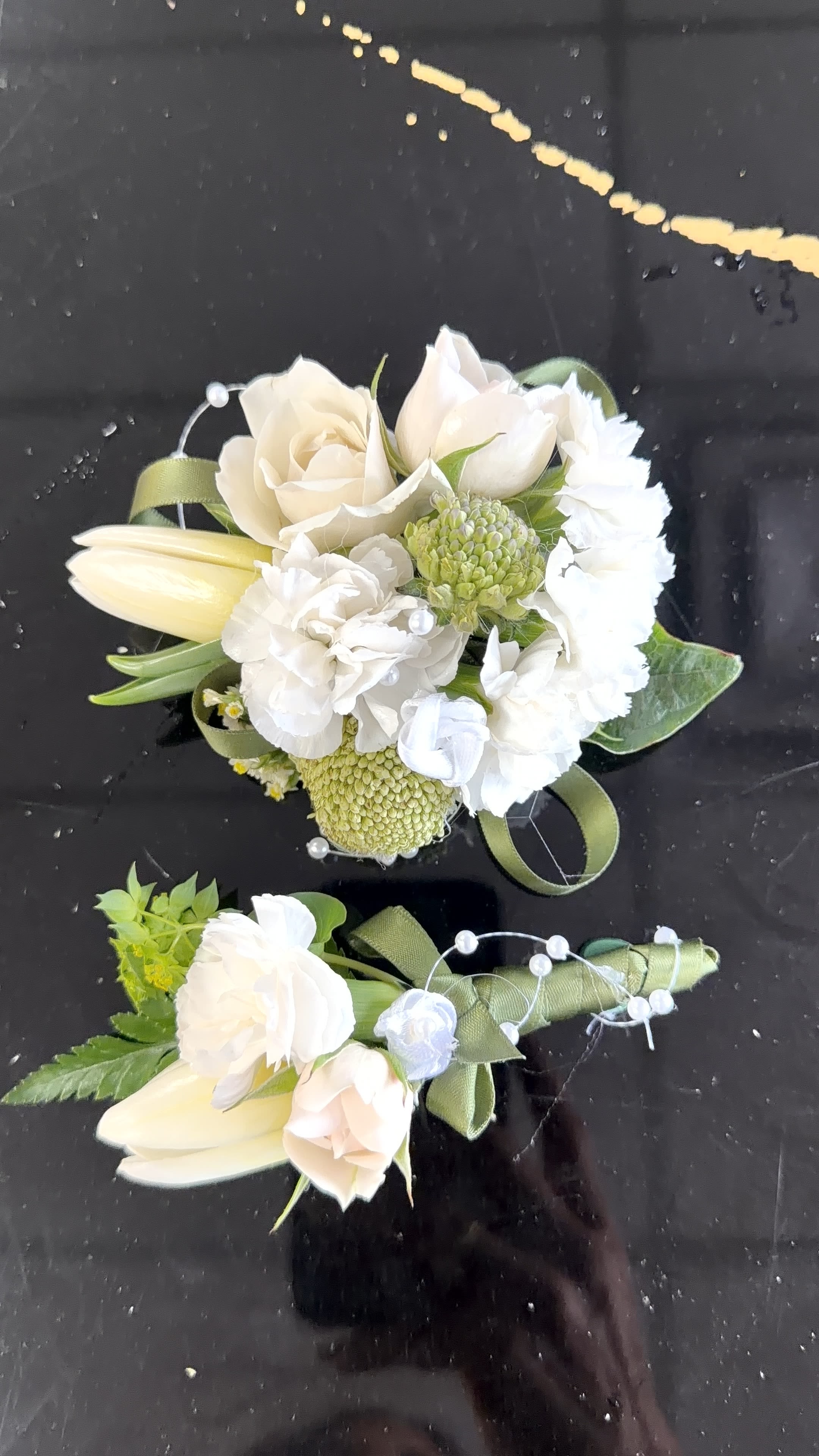 White Corsage and white Botonier  - Delicate cream-and-white blooms shaped into a classic white corsage and matching boutonniere create an elegant finishing touch for weddings, proms, or any formal event. The soft neutral palette-white, peach, and fresh green-gives the recipient a timeless, refined look that reads as both romantic and understated; the person wearing it will feel polished and perfectly dressed for the occasion.  Each piece combines rose-like blooms, tulip buds, ruffled carnations, and small chrysanthemums for a varied texture that's soft to the eye. Arranged at a compact scale, the corsages feature neat green ribbon loops and decorative pearl picks that catch the light and add a subtle, formal sparkle. The boutonniere mirrors the same flowers and accents so couples or pairs look coordinated without being matchy.  Ideal for bridesmaids, prom dates, parents, or honored guests, this set signals thoughtful attention to detail and complements ivory, blush, or green palettes. The compact size is designed to sit comfortably on lapels and wrists; keep them cool and mist lightly if needed before wear to preserve freshness.