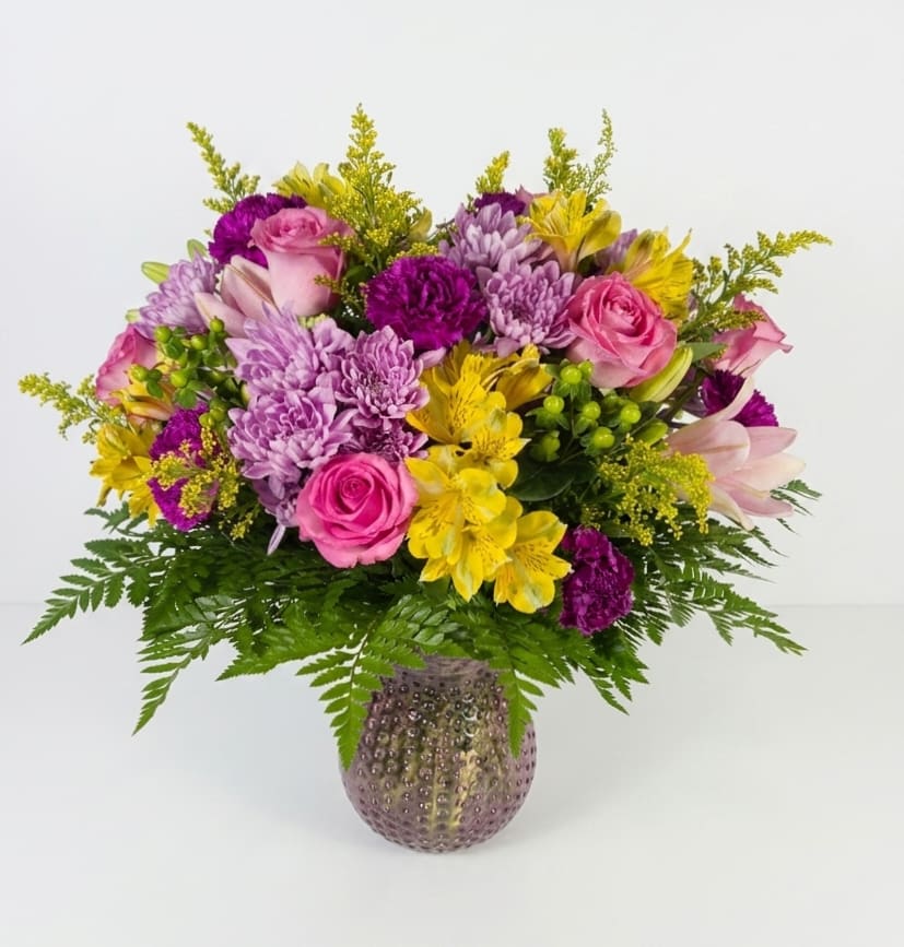 Sunshine Meadow  - A lively bouquet of mixed color roses, alstromeria, chrysanthemums and airy filler that feels like a sunlit meadow perfect as a birthday bouquet, a cheerful thank-you, or an everyday gift to brighten someone's day. The mix of pink roses, purple chrysanthemums and yellow alstromeria creates a warm, celebratory contrast that will make the person you're thinking of feel seen and uplifted.  Arranged in a textured pink glass vase, this full, rounded design stands about 18" tall and brings both color and texture to any room. The bright yellow blooms balance the cooler pink and lavender tones for a harmonious, festive look; the textured vase adds an elegant, decorative touch that works equally well on a mantel, desk, or dining table.