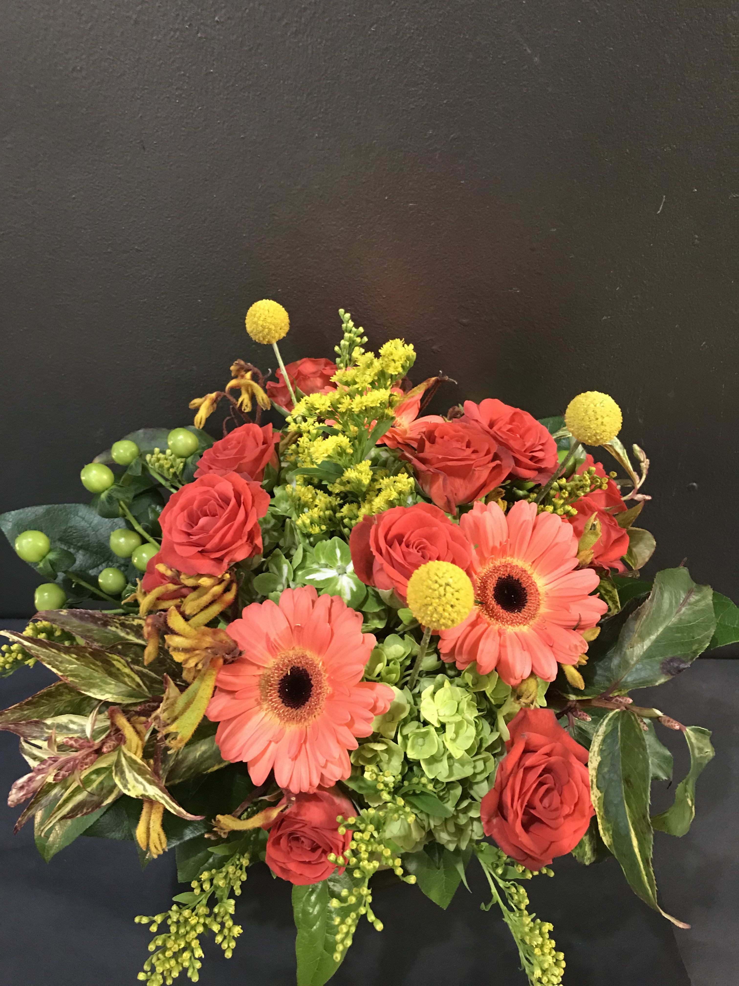 Compact arrangement of coral gerbera daisies, red roses, green hydrangea, and yellow accent flowers.