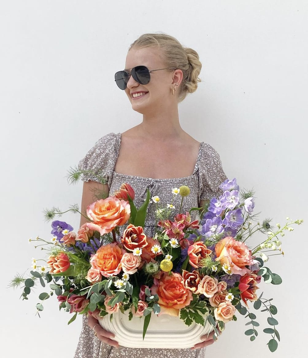 The Elise - The Elise embodies modern elegance with a vibrant mix of delicate blooms. Free spirit roses and peach spray roses form a warm, inviting base, while fluffy light purple delphinium and bright pink alstroemeria add lively pops of color. Pink double tulips and soft blue nigella lend a whimsical touch, beautifully complemented by cheerful feverfew and fresh eucalyptus. Arranged in a long, low geometric vessel, the Elise brings a sophisticated, contemporary charm to any setting.