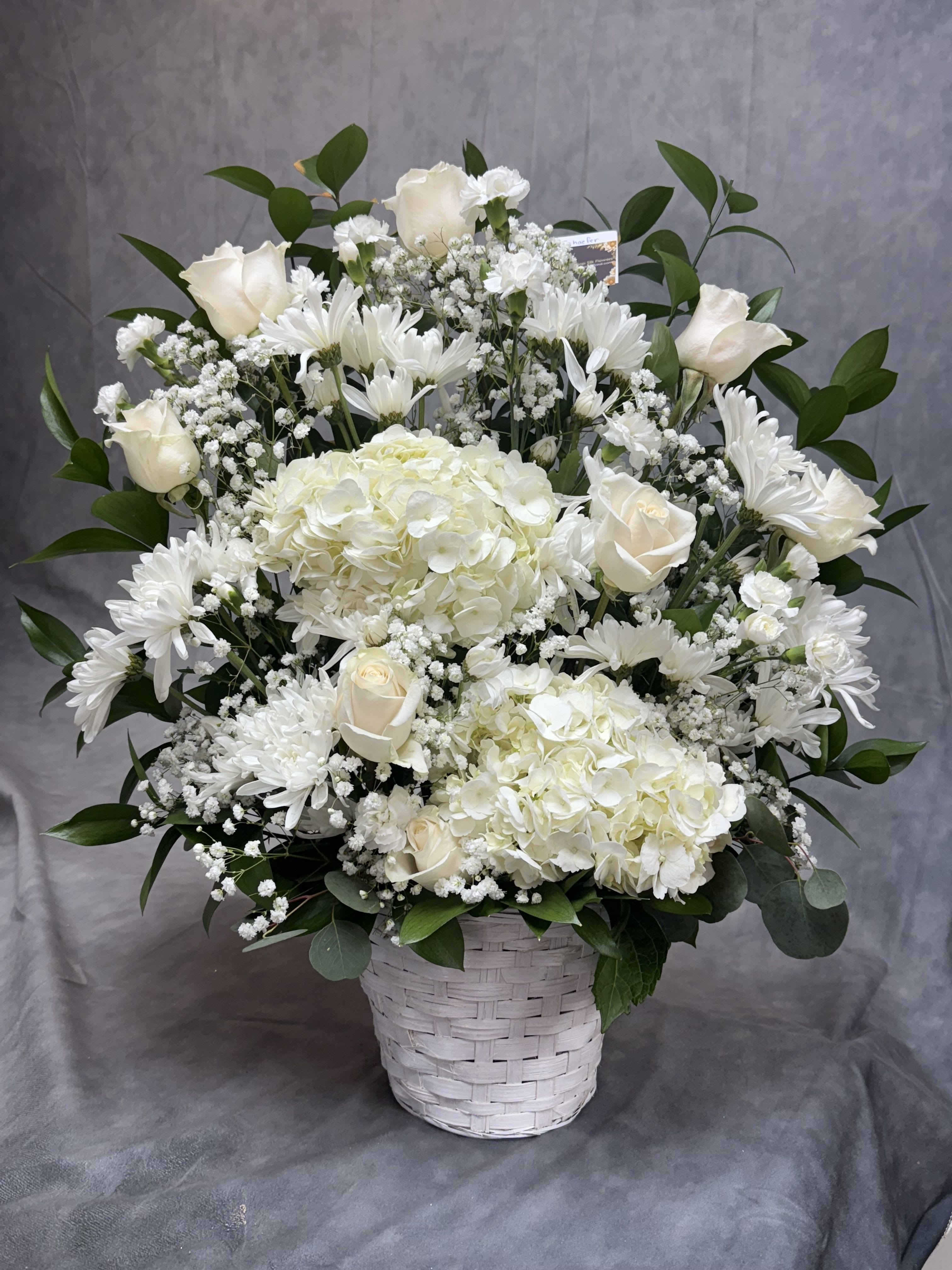 White floral arrangement in a woven basket with roses, hydrangeas, and daisies