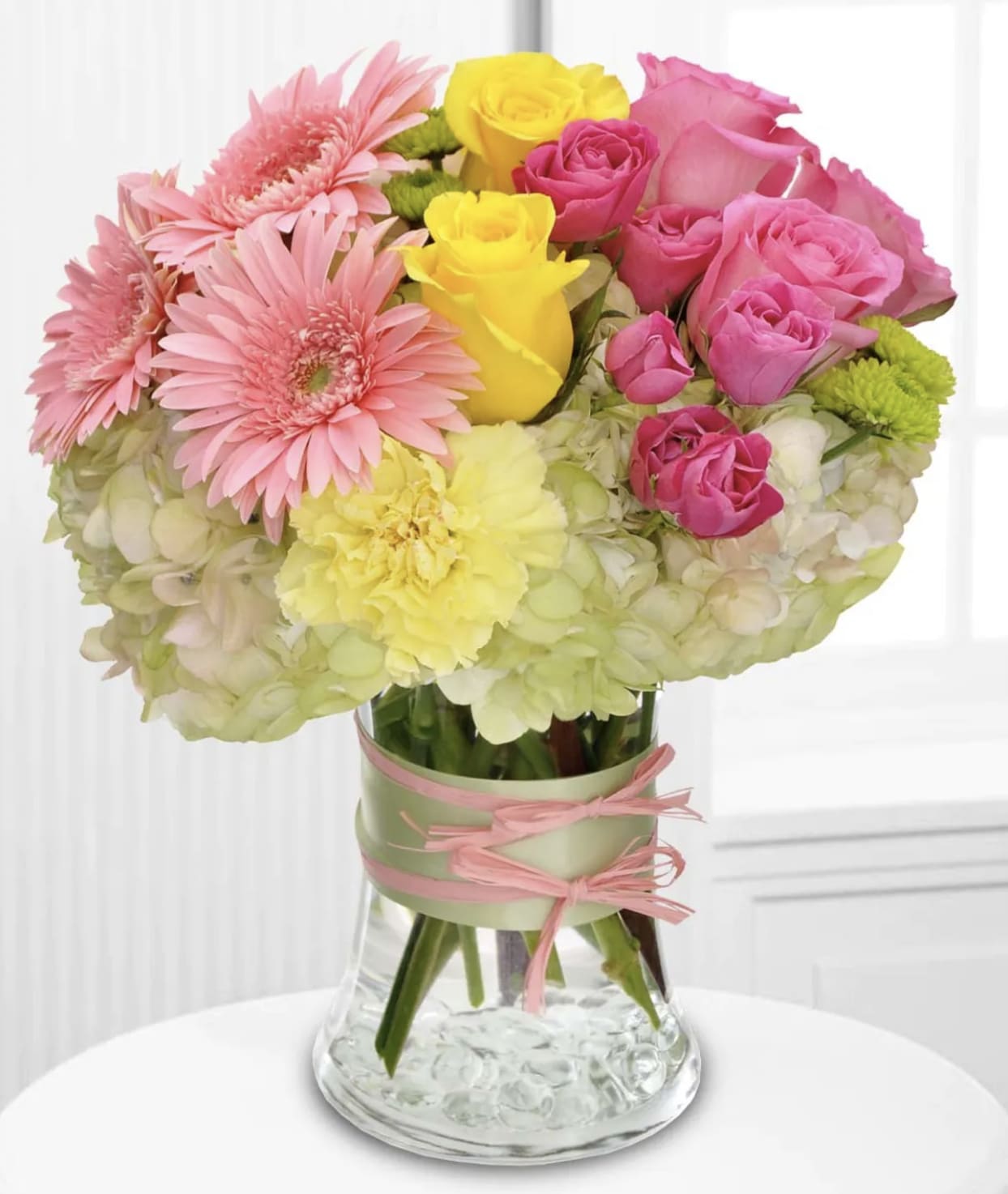 Fashionista Blooms - This arrangement would be perfect for any girl with an eye for style. It's a must-have for fashionistas everywhere.  Gorgeous green hydrangea, yellow and light pink roses, pink spray roses and mini gerberas, light yellow carnations and green button spray chrysanthemums are delivered in a pretty gathering vase. Not just any vase, of course, this one's accessorized with a chartreuse taffeta ribbon and pink raffia.  Approximately 10" W x 11" H  Orientation: All-Around      As Shown : T155-1A     Deluxe : T155-1B     Premium : T155-1C  