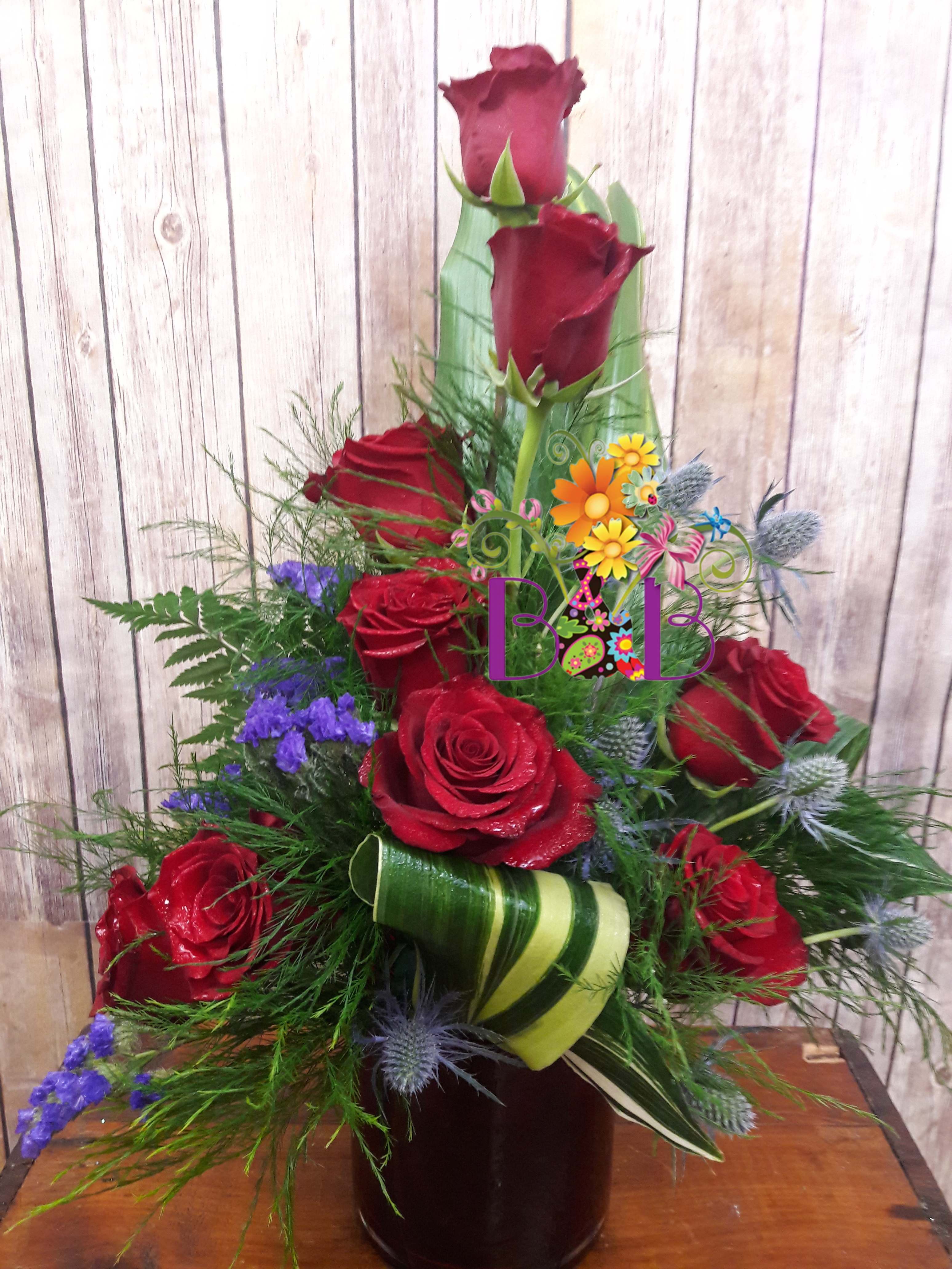 Red roses arranged in a tall vase with purple filler flowers and green ribbon accents