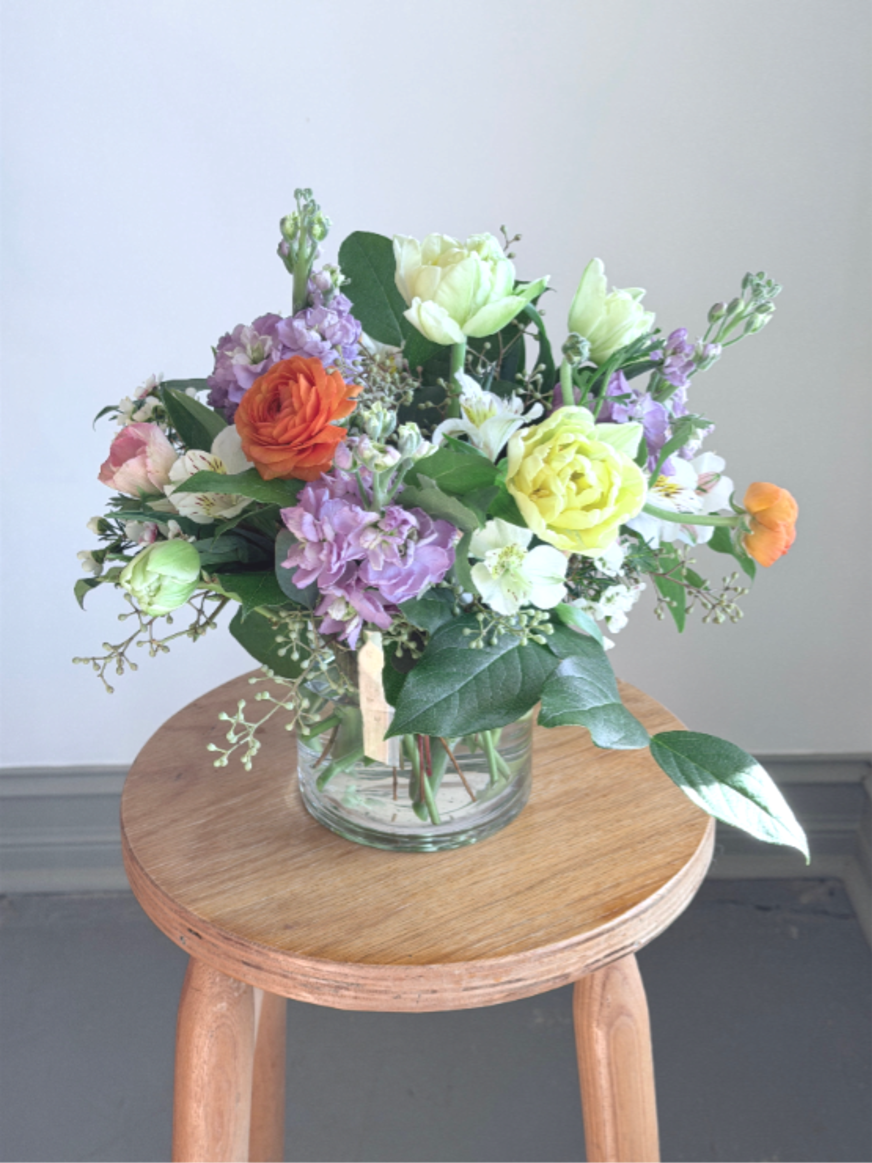 Mixed pastel flower arrangement with yellow roses and purple blooms in a clear glass vase on a wooden stool.