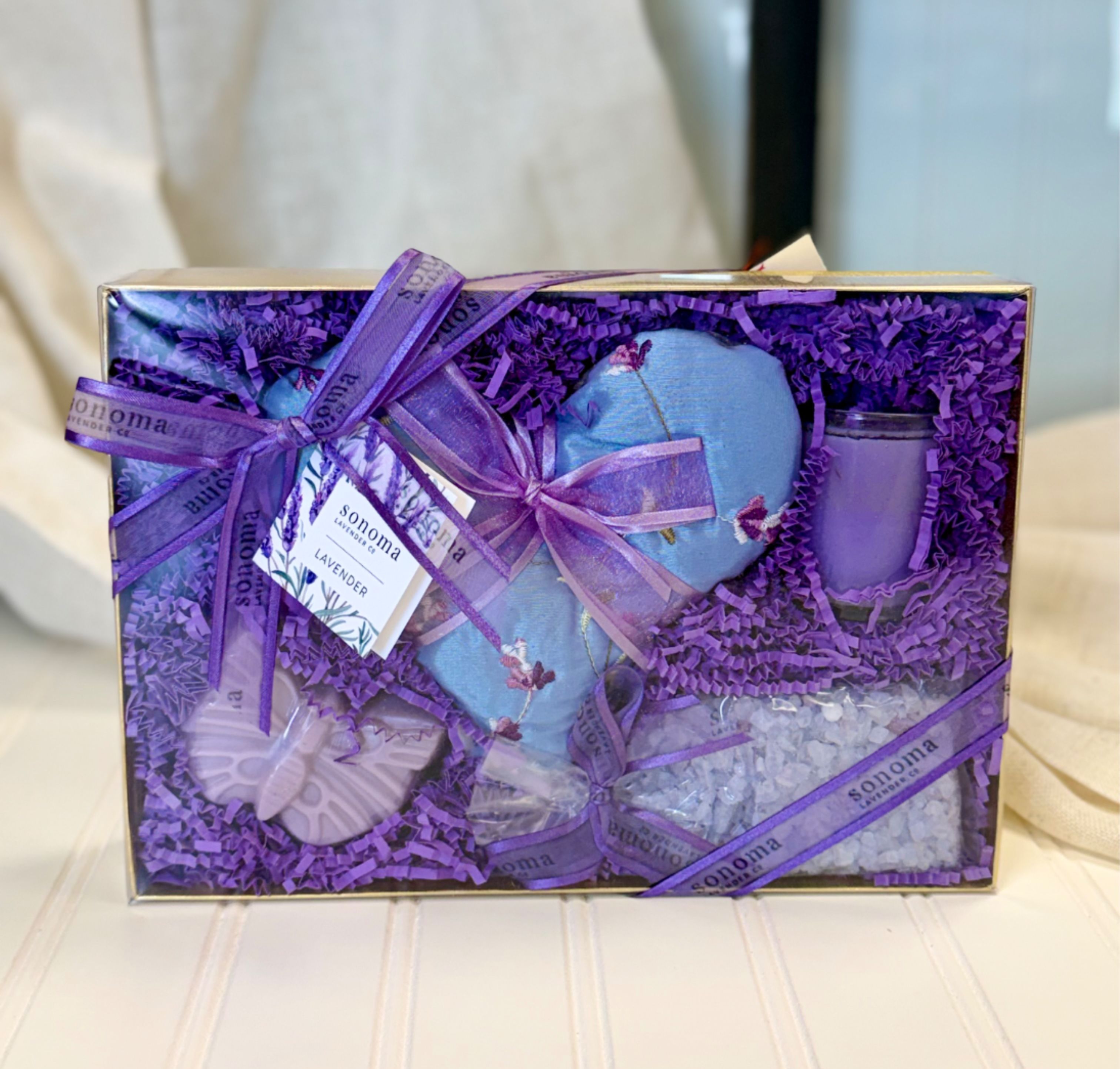 Lavender Gift Set - Soft lavender and serene blues set the tone for a calming self-care gift-perfect as a relaxation present or a thoughtful birthday or thank-you gift. This lavender gift set is presented in a rectangular gift box tied with a sheer ribbon, making it ready to hand to the person you want to soothe and pamper. The color story of purple, blue, and white reads peaceful and restorative, ideal for someone needing a moment of quiet or a little at-home spa escape.  Inside the box, purple crinkle filler cushions a coordinated collection: a heart-shaped lavender sachet, a scented candle, bath salts, and a soap-like bar, all styled in soft purple and blue tones. The neat presentation and ribbon ties make the set feel polished and intentional, while the sachet and bath items deliver layered fragrance and tactile comfort. Let this box be the thing that helps someone unwind after a long week-the kind of gift that says "I'm thinking of you" without needing words.