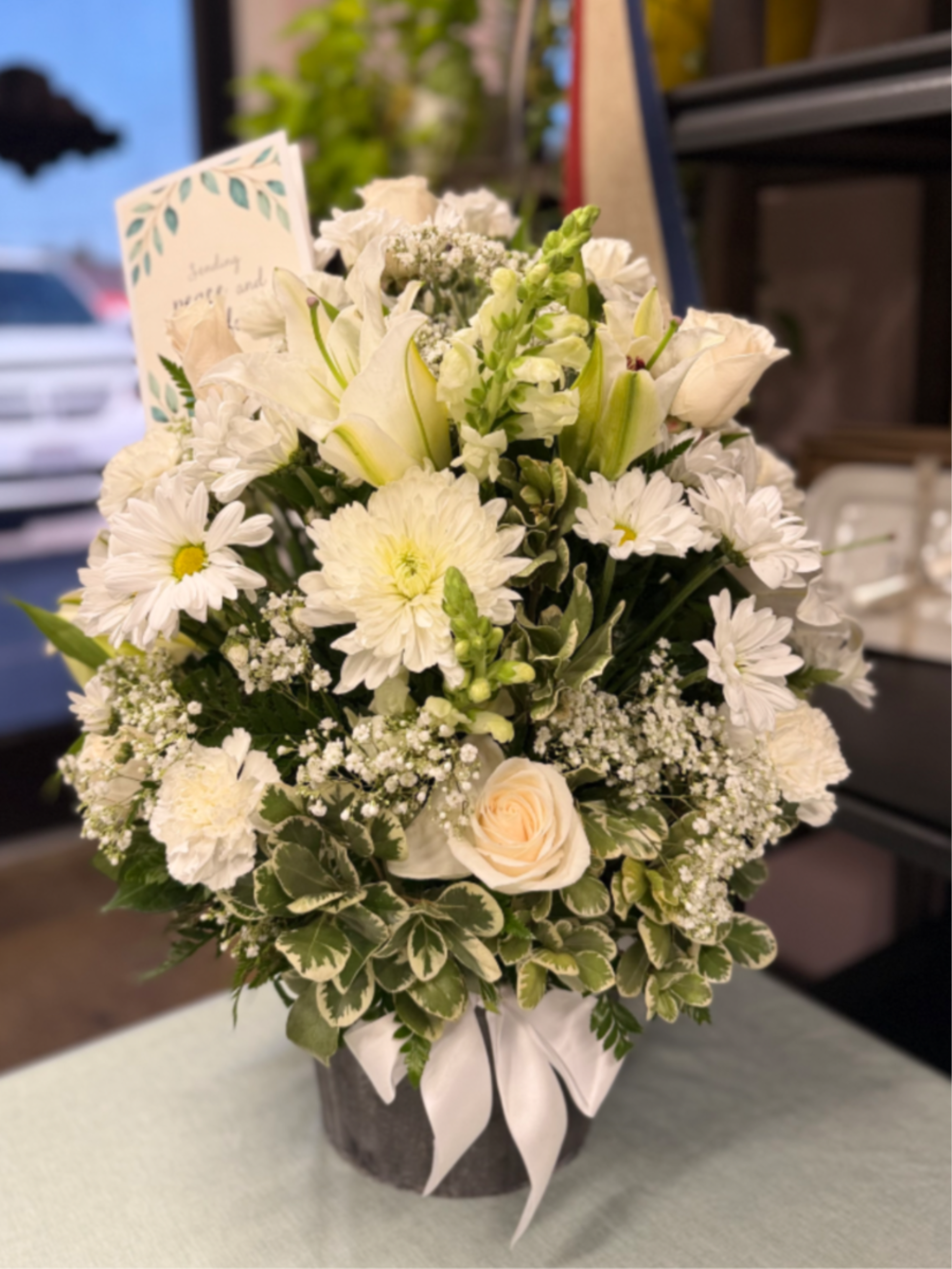 Memorial Basket - This elegant basket arrangement features a serene blend of white lilies, carnations, roses, and mums, beautifully accented with delicate baby’s breath. Designed with lush greenery, it offers a timeless expression of peace, respect, and remembrance—an ideal tribute to honor a loved one’s memory. Size: Medium approximately 22 inches tall    