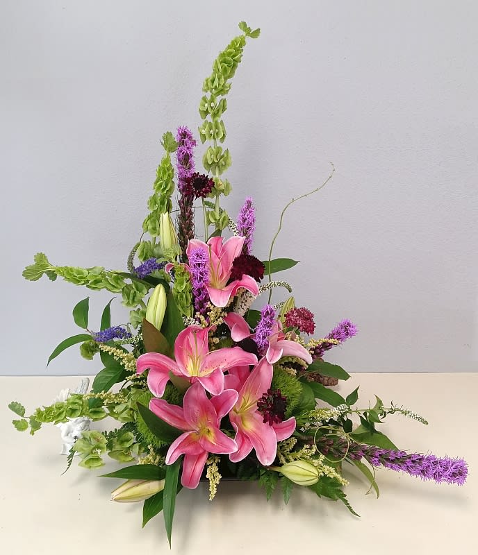 Pink lilies arranged with tall green and purple flowers in a low container
