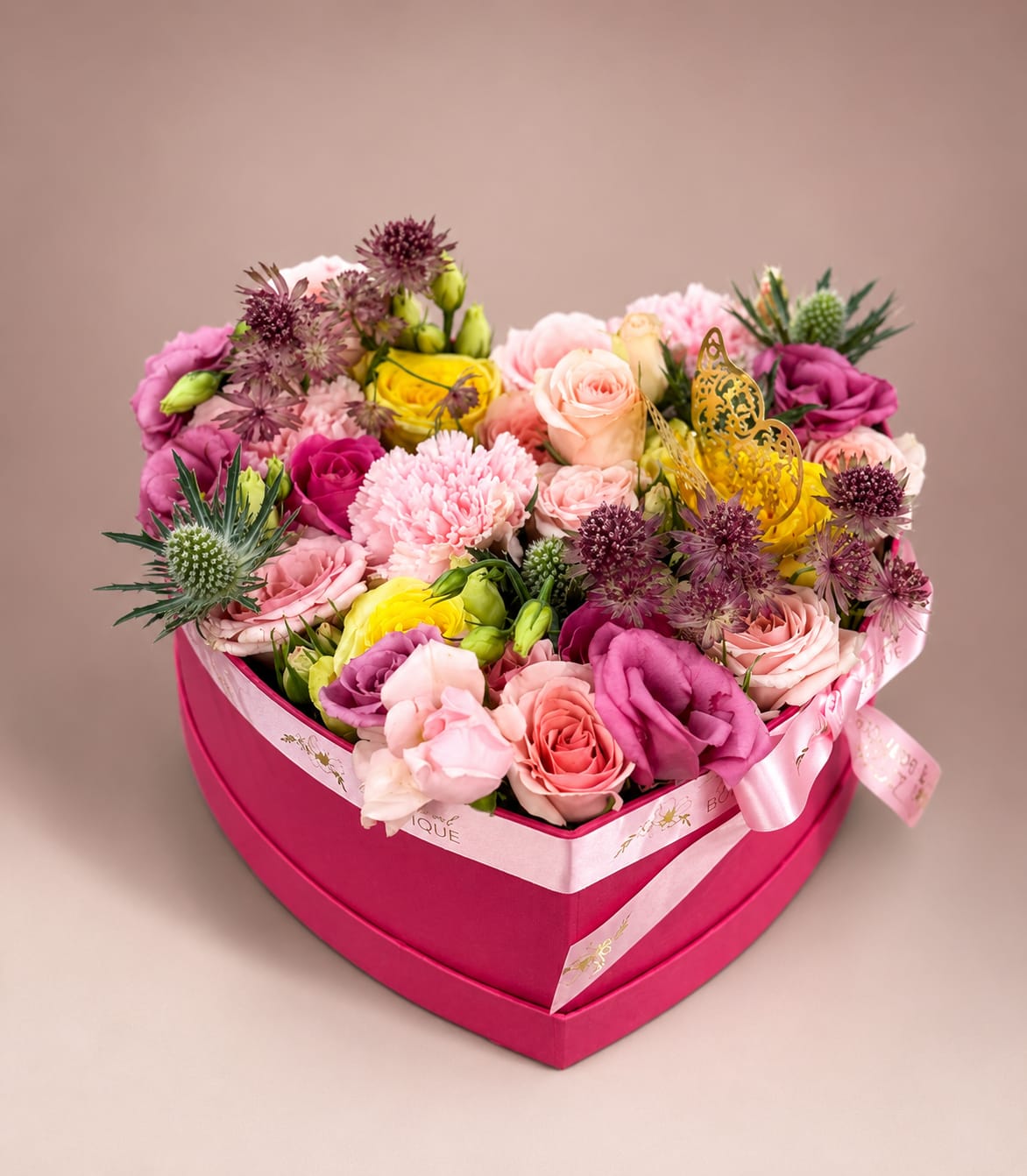 Spring Heart Box  - A bright heart-shaped pink box overflowing with roses, carnations, lisianthus and other petite accent blooms makes this flower arrangement a cheerful everyday gift that reads as romantic and playful - perfect for a birthday, anniversary, or just because. The mixed-color palette of hot pink, blush, sunny yellow and soft lavender-purple creates a layered, sunlit look that will make the person you're thinking of feel truly seen and celebrated.  Tightly packed and layered for a full, abundant silhouette, the blooms sit snugly in the heart box and are finished with a satin ribbon and a decorative butterfly pick for a polished, gift-ready presentation. Compact but generous in appearance, this arrangement is ideal for a bedside table, desk, or mantle and sends a warm, joyful message the moment it arrives.