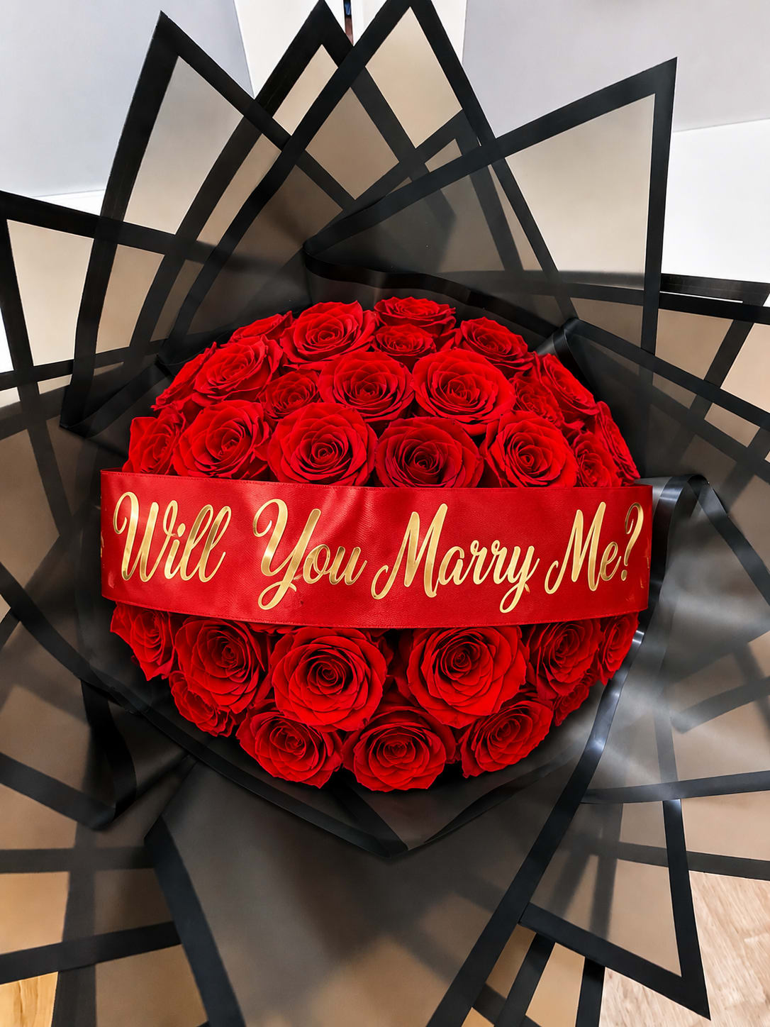 Will You Marry Bouqet  - Create an unforgettable moment with our Signature Proposal Bouquet, a luxurious hand-tied arrangement of premium long-stem roses wrapped in elegant premium wrapping for a sophisticated presentation. Finished with a personalized satin ribbon, this bouquet is perfect for proposals, anniversaries, or any romantic milestone.  Customize your ribbon with messages such as “Will You Marry Me?”, “Will You Be My Girlfriend?”, or another special phrase to make your gift truly one of a kind.  Rose colors can also be customized upon request, allowing you to create a bouquet that reflects your loved one’s favorite colors or your unique vision.  Features:  - Premium long-stem roses - Luxury Premium Wrapping - Personalized satin ribbon with your custom message (Available with messages like “Will You Marry Me?” or “Will You Be My Girlfriend?”) - Rose colors can be adjusted upon request   *** Each custom proposal bouquet is handcrafted to order. To ensure the highest quality and allow time for personalization, orders must be placed a minimum of one day (24 hours) before your requested pickup or delivery date.