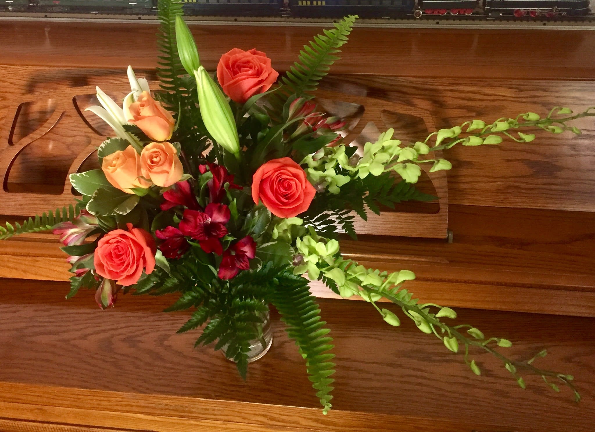 Mixed arrangement of orange roses, white lilies, red blooms, and green orchids in a clear glass vase