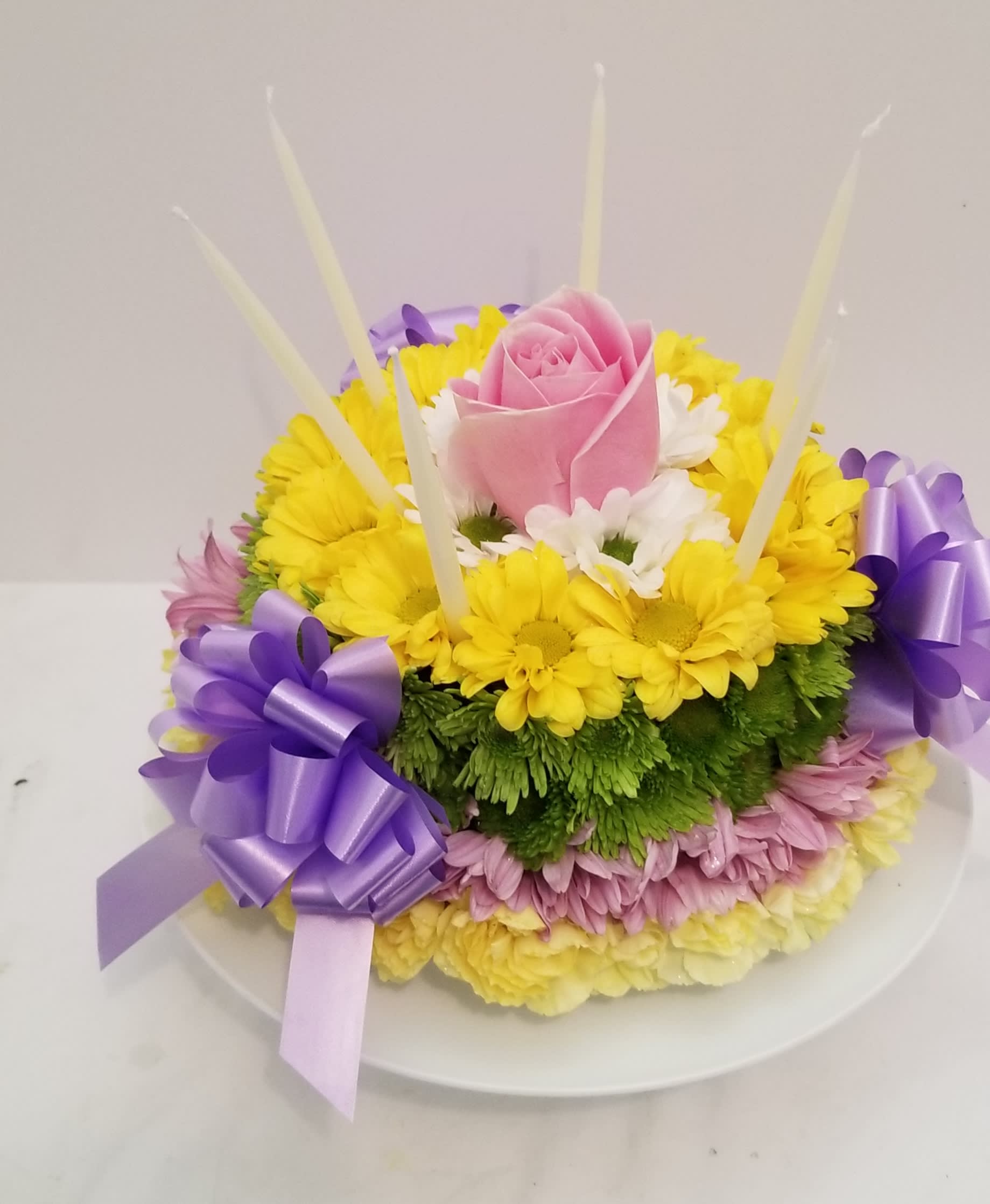 Round floral cake of yellow and pink blooms with a pink rose, purple bows, and tall white candles
