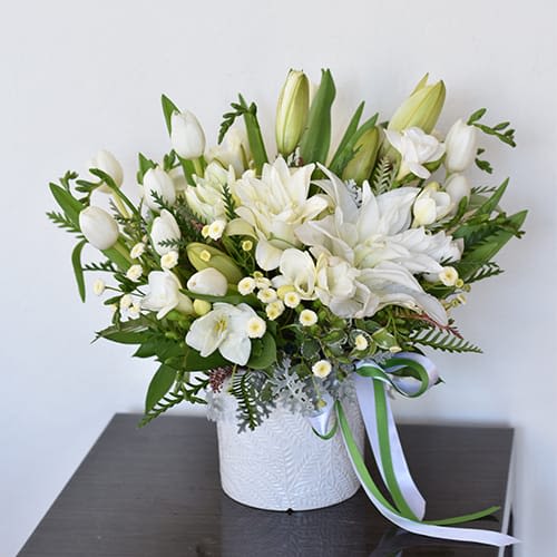 See You Again Boquet - "See You Again Bouquet" arrangement tenderly combines the gentle allure of white tulips with the solemn grace of white lilies, offering solace and comfort during times of loss.  At its heart, pristine white lilies stand tall, their serene blooms symbolizing purity and the promise of peace beyond grief. Their delicate fragrance whispers of remembrance, soothing sorrow-filled hearts with each delicate petal.  Encircling the lilies are clusters of white tulips, their soft, velvety petals echoing the tender memories shared with the departed. Each tulip, with its understated elegance, reflects the cherished moments and enduring love woven into the tapestry of friendship.  Interwoven amidst the blooms, verdant foliage adds a touch of natural beauty, symbolizing the eternal cycle of life and offering a glimpse of hope amidst the darkness of loss. Their lush greenery serves as a reminder that though parted, the spirit of the departed lives on in the hearts of those who hold them dear.  Together, the white tulips and lilies create a serene tableau of comfort and support, honoring the memory of a cherished friend while offering solace and strength to those left behind.