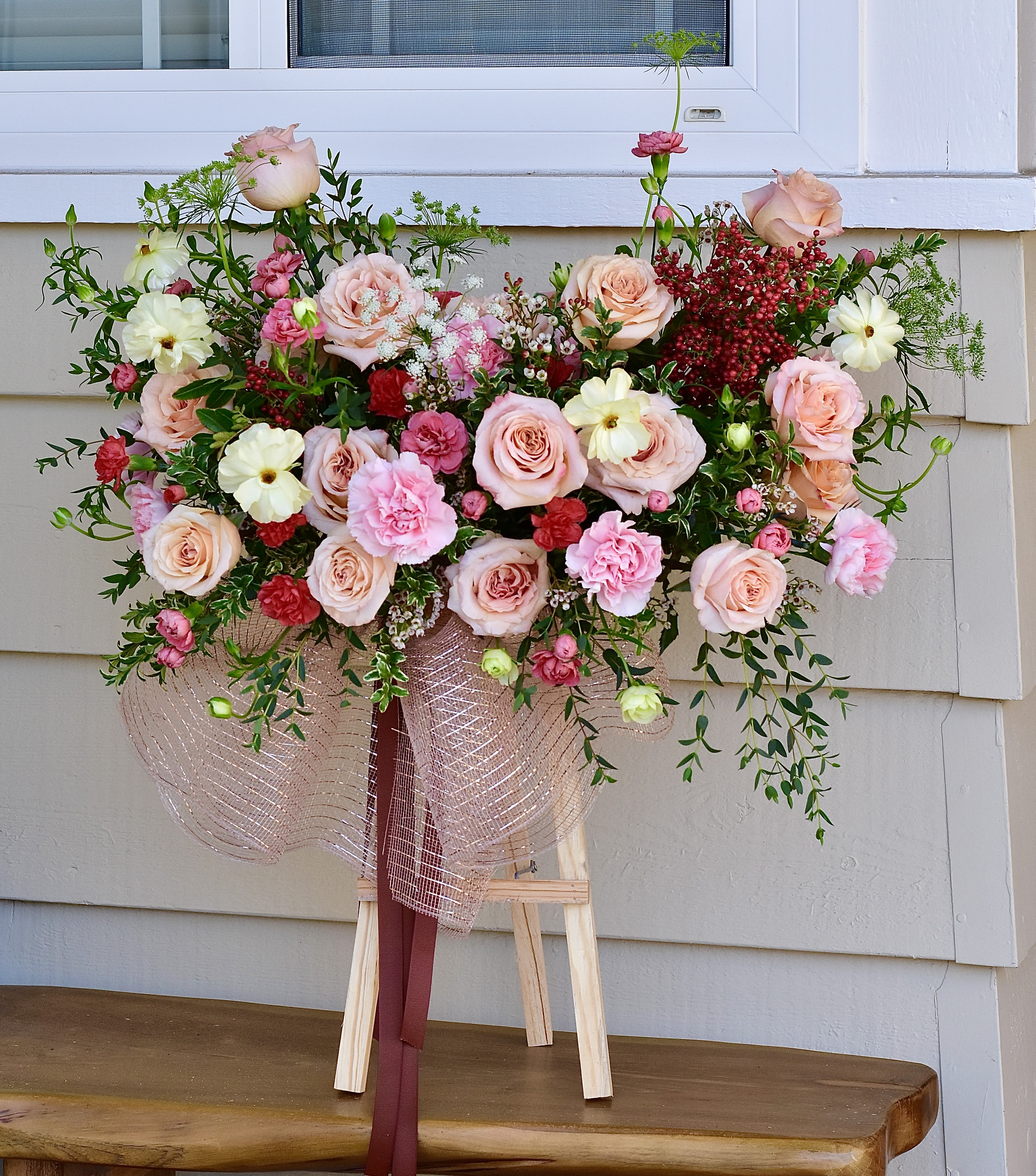 Grand Opening Table Easel - ake a bold statement on opening day! Our Grand Opening Table Easel is designed to capture the excitement of a new chapter. This vibrant arrangement showcases two dozen radiant roses accented by a dozen carnations and charming mini-carnations for a full, voluminous look.  The whimsical touch of Queen Anne’s Lace provides a delicate, "wildflower" contrast to the structured beauty of the roses, while lush green fillers add depth and freshness. It’s a gift of prestige and joy, perfect for any storefront or reception gala.