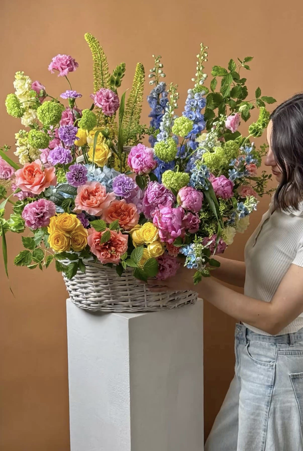 Special occasion Basket - A tall, joyful mixed flower arrangement in a white wicker basket - this large celebration bouquet reads as a festive birthday or congratulations gift that immediately lifts the room. Bright pink roses and coral accents mingle with sunny yellow chrysanthemums, sky-blue delphinium, and lime-green touches to create a layered, garden-like scene with strong vertical movement that catches the eye.  Rounded roses and carnations contrast with spiky delphinium stems for lively texture and a playful silhouette that feels both abundant and airy. The design's generous scale and mixed color palette make it perfect for spring events, milestone celebrations, or as a cheerful centerpiece to mark someone's big day; the pedestal display adds formality while keeping the overall impression light and celebratory.