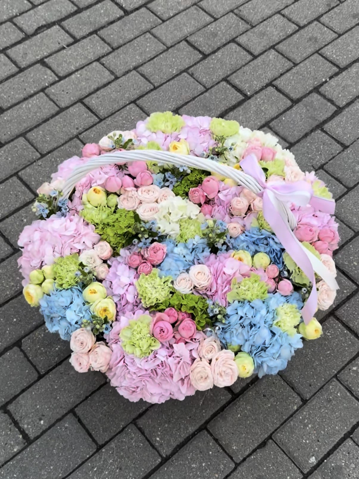 Basket of Joy - A lush, pastel-filled gift for spring celebrations, this flower arrangement is a cheerful basket of joy perfect for birthdays, Mother's Day, or to say you're thinking of someone special. The soft mix of pink, blue, purple and green blooms creates a romantic yet lighthearted feel that will make the recipient feel genuinely celebrated-an ideal choice when you want to brighten a room and lift someone's spirits.  Densely arranged in a white wicker basket, the composition pairs roses, hydrangeas, carnations and delicate filler blossoms in layered tones of pink, lavender, cream and powder blue. A pale lavender ribbon tied to the handle gives it a finished, gift-ready look; the round, abundant silhouette reads like a floral hug and fills tabletops or entryways with texture and color. The basket presentation makes it easy to place without a vase while still feeling elegant and thoughtful.  Designed for moments that call for warmth and celebration, this arrangement suits mothers, friends, new parents or anyone who appreciates soft, romantic florals. Sizes are generously full to create that overflowing, decorative impression-treat the person you're thinking of to an arrangement that feels personal and joyful. For best freshness, keep in a cool spot away from direct sun and follow any care slip included with your delivery.