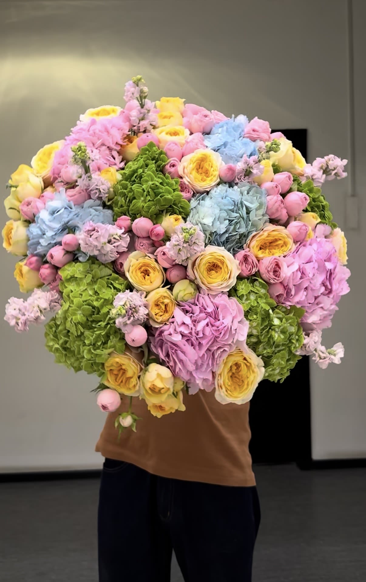 Dopamine flowers signature wrap - A lush, oversized hand-tied bouquet that instantly lifts spirits with a playful pastel palette - perfect as a birthday bouquet, anniversary flowers, or a cheerful thank-you arrangement. This garden-style wrap combines the rounded volume of hydrangeas with romantic roses and the delicate layers of ranunculus, accented by tall, fragrant stock for a soft but lively look your recipient will remember.  Soft pinks, buttery yellows, sky blues, and bright greens mingle in a densely packed, rounded silhouette that feels abundant and thoughtfully layered. The mix of large hydrangea heads, full roses, and textured ranunculus creates depth and a tactile, petal-rich experience, while the stock adds gentle vertical interest. Sized as a large hand-held bouquet, it makes a bold gifting statement without being formal.  Give this to someone who deserves to feel genuinely celebrated - it reads as joyful, romantic, and refreshingly modern for spring occasions or everyday celebrations. Hand-tied and wrapped for easy carrying, enjoy fresh-cut care by keeping stems in clean water and trimming an inch every few days to prolong bloom life.