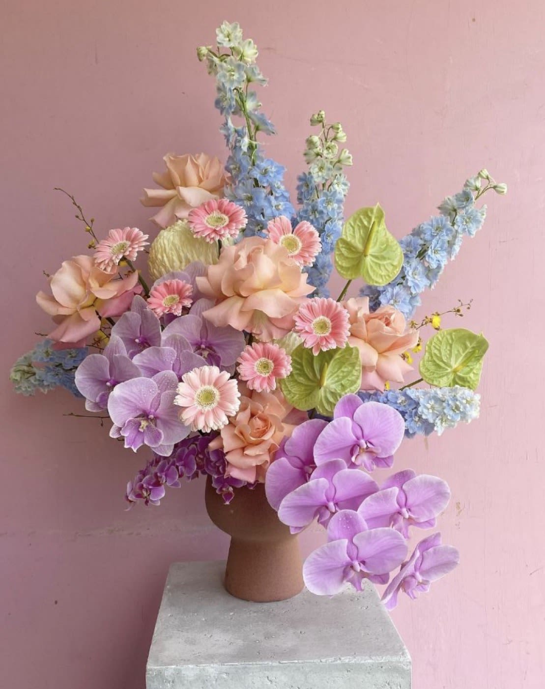 Colorful centerpiece  - Lavender phalaenopsis orchids set a dreamy tone in this colorful centerpiece, perfect as a birthday bouquet or romantic table arrangement. The mix of pink gerbera daisies and peach roses with tall blue delphinium-like spikes gives the piece a whimsical, celebratory feel-the kind of floral gift that makes the person you're thinking of feel adored and noticed.  This full, asymmetrical arrangement is composed in a low ceramic vase, combining large lavender and purple orchids, peach roses, pink gerbera daisies, bright green anthurium blooms, and airy blue spikes for height and movement. Several stems reach upward and outward to create a dramatic silhouette; textures shift from glossy anthurium leaves to soft rose petals for visual depth. Ideal for a centerpiece on a dining or event table, it arrives ready to display-no extra arranging required-and will bring pastel romance and lively color to any celebration.