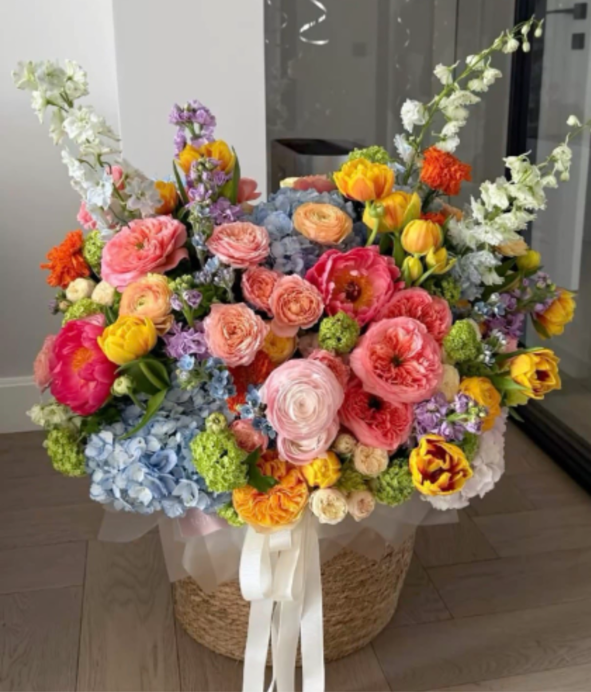 Happy Birthday Basket  - Layered pink and coral roses meet  pale blue hydrangeas in a large, garden‑like birthday basket that feels both celebratory and romantic - a perfect birthday bouquet or Mother's Day gift. This Happy Birthday Basket is designed to make the person you're thinking of feel truly seen: abundant, rounded, and bursting with pastel charm brightened by vivid orange and magenta accents.  The arrangement is densely packed in a woven basket, tied with a long white ribbon and finished in a translucent clear wrap for a gift-ready presentation. You'll find pink and peach ranunculus, stock and white branching blooms, purple accent flowers, roses, and clusters of hydrangea arranged for a layered, textured look that reads lively from every angle. Colors include soft blues, pinks, whites, purples, and cheerful yellows to create a balanced, uplifting palette.  Ideal for birthdays, anniversaries, or as a festive "thinking of you" surprise, this arrangement suits dining tables, entryway displays, or as the centerpiece at a small celebration. The woven basket and sheer wrap make it easy to present straight away; minimal care-fresh water and a cool spot-will keep the blooms looking their best through the special day and beyond.