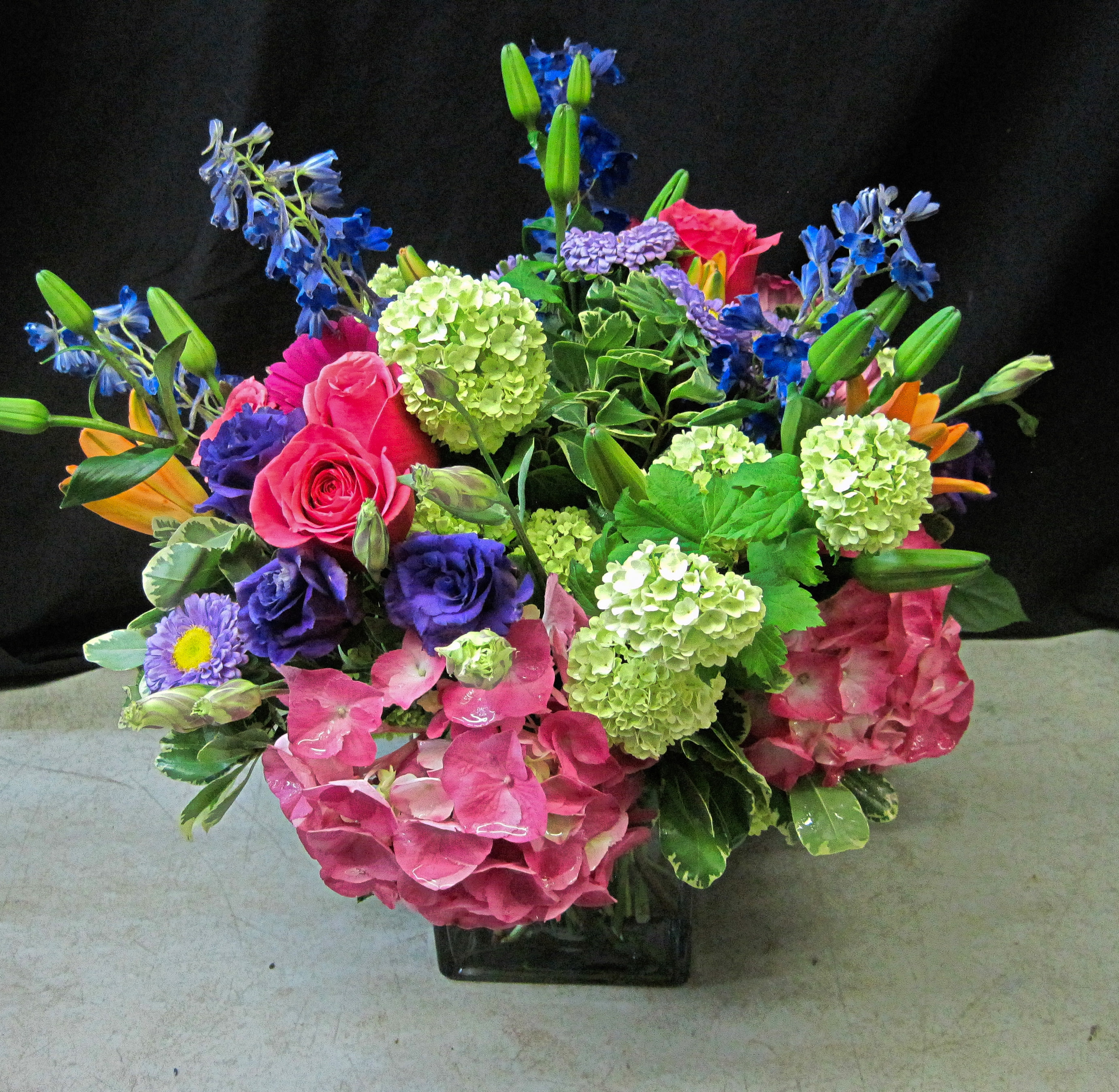 Colorful arrangement of pink hydrangeas, roses, green blooms and mixed purple and blue flowers in a glass cube vase