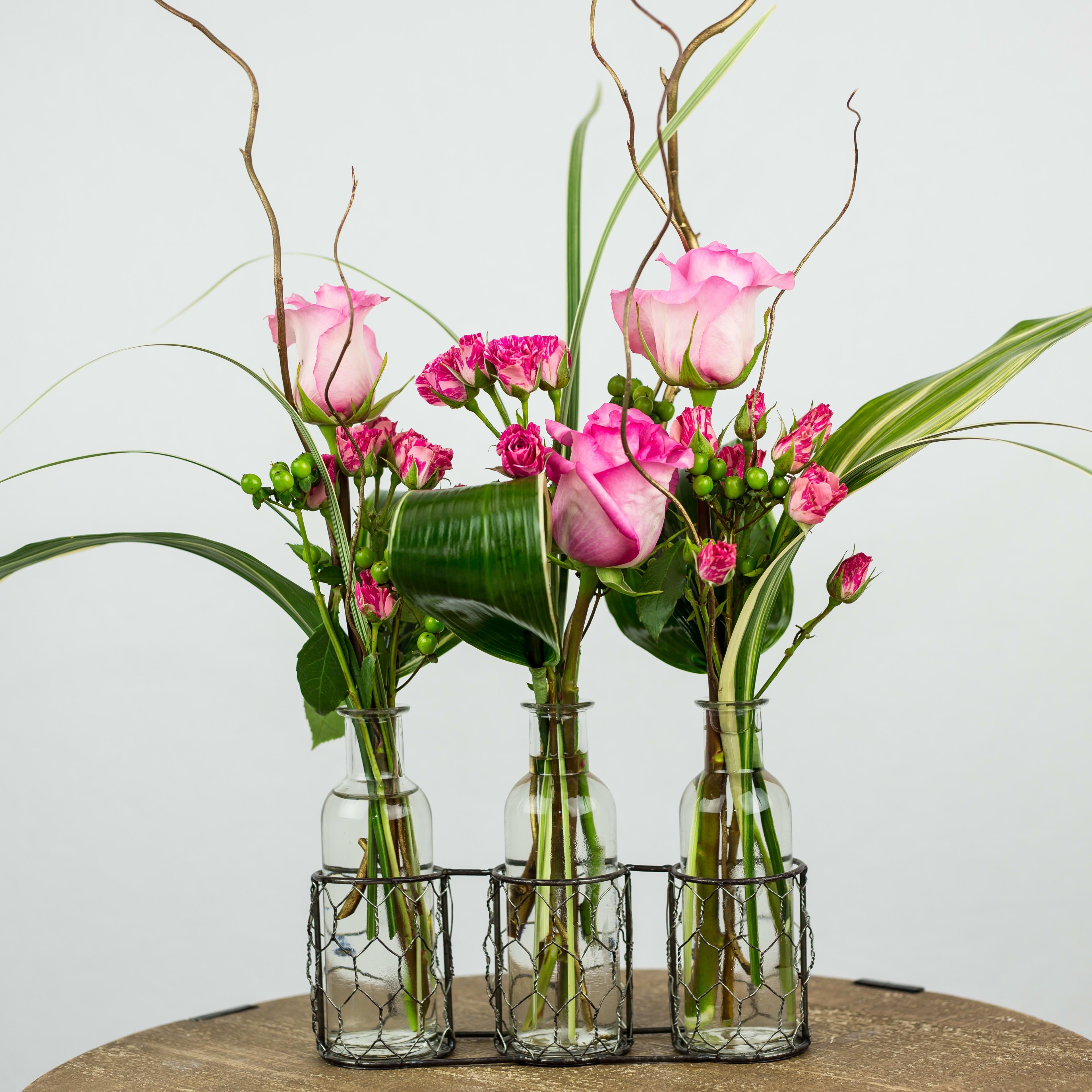 Three small glass vases hold pink roses and carnations with tall decorative branches.