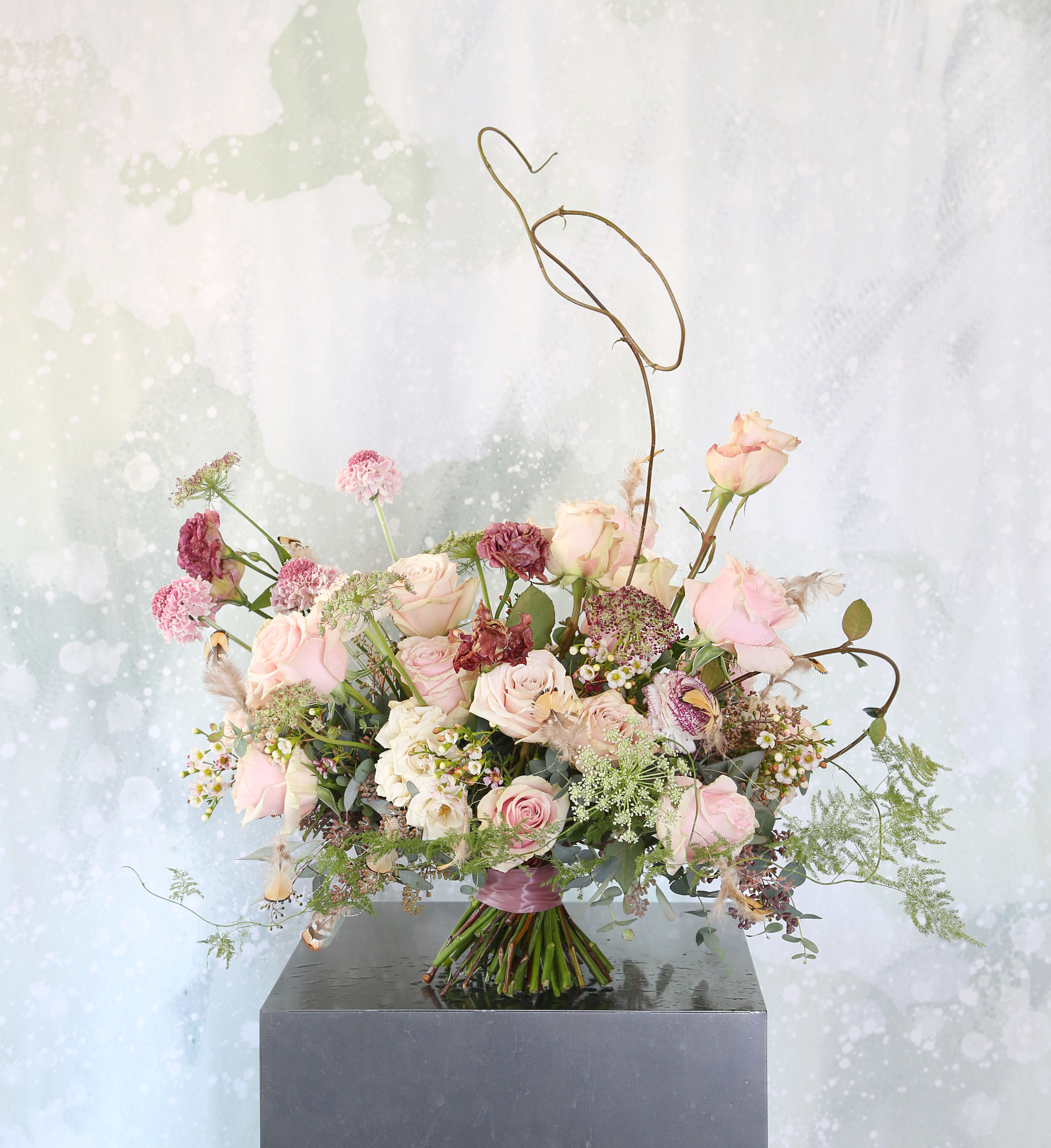 Loose hand-tied bouquet of blush and mauve roses with airy accent flowers and sculptural wire detail.