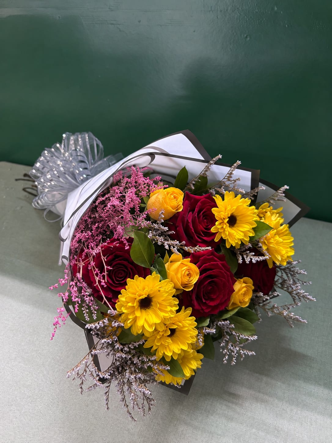 Sunset Berry Bouquet -  A vibrant hand-tied bouquet featuring rich burgundy roses, sunny yellow blooms, soft pink accents, and fresh greenery. Wrapped in crisp white and black paper, this cheerful arrangement is ready to grab, gift, and brighten someone’s day.