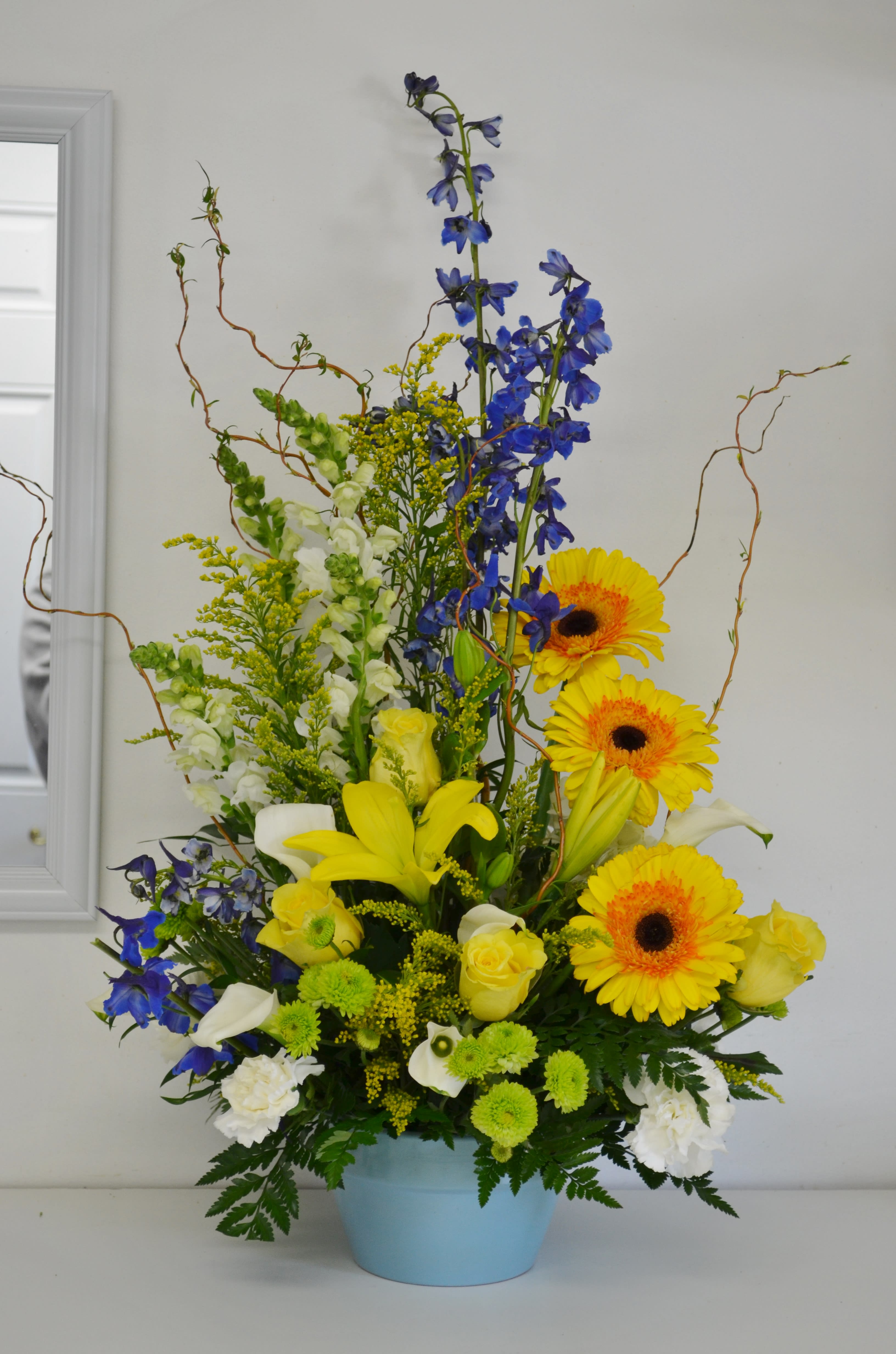 Tall mixed bouquet in a blue pot with yellow gerberas, blue delphinium, and white lilies